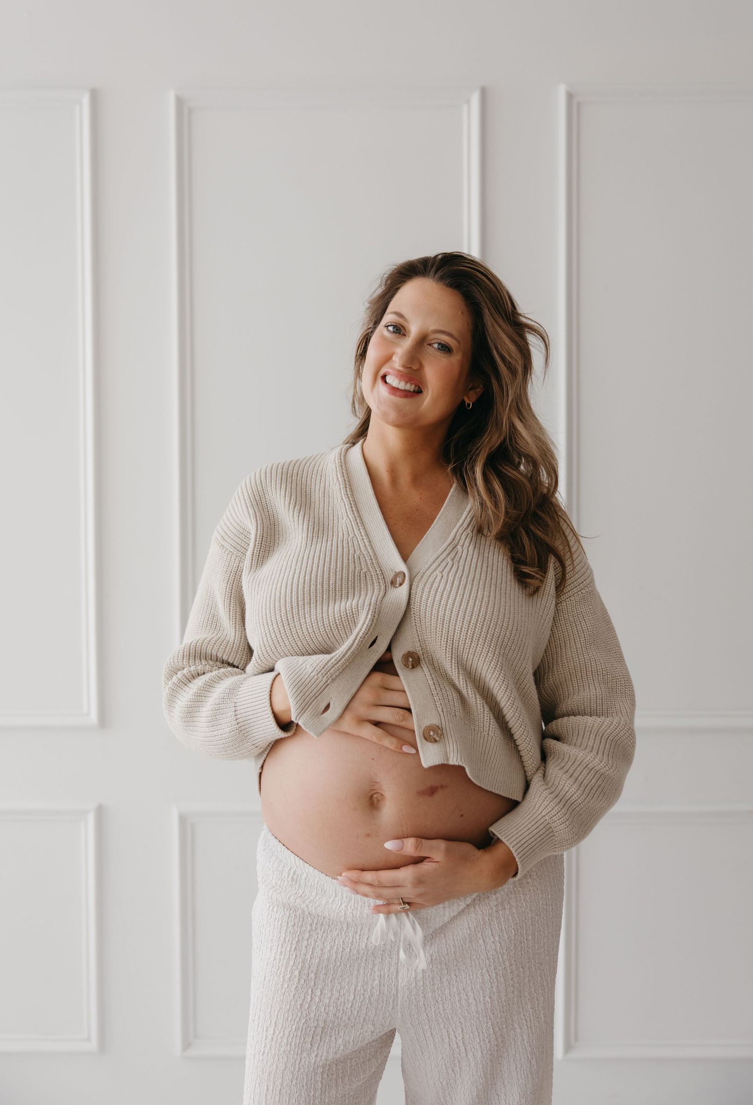 Smiling pregnant woman in beige ribbed cardigan and pants cradling baby bump against white paneled wall.