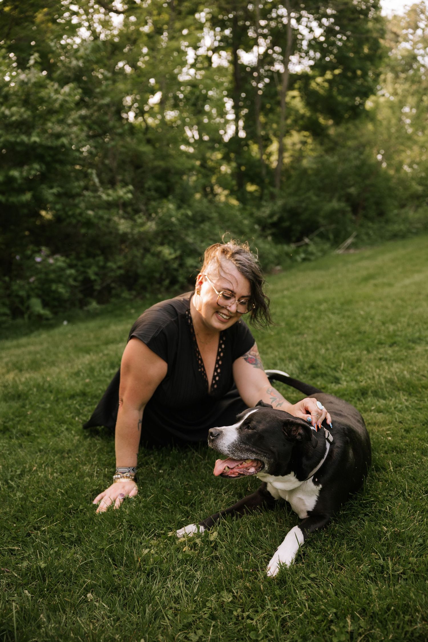 Smiling woman with glasses pets a black and white dog while sitting on green grass.