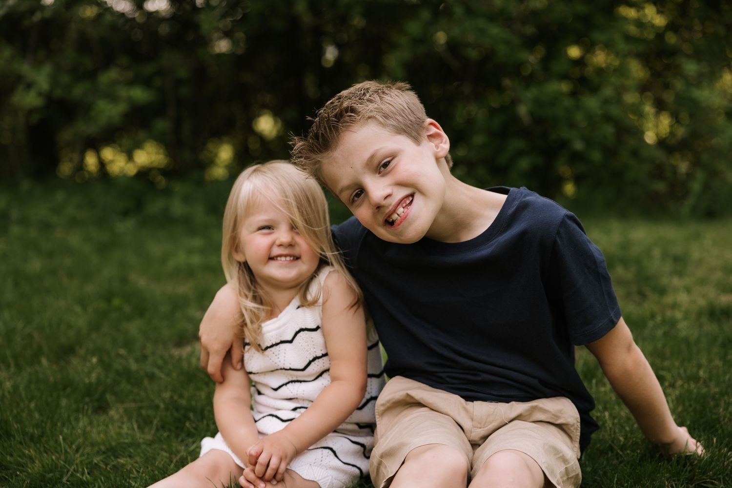 Two happy children, a young girl and older boy, sit together smiling on green grass in a sunny outdoor setting.