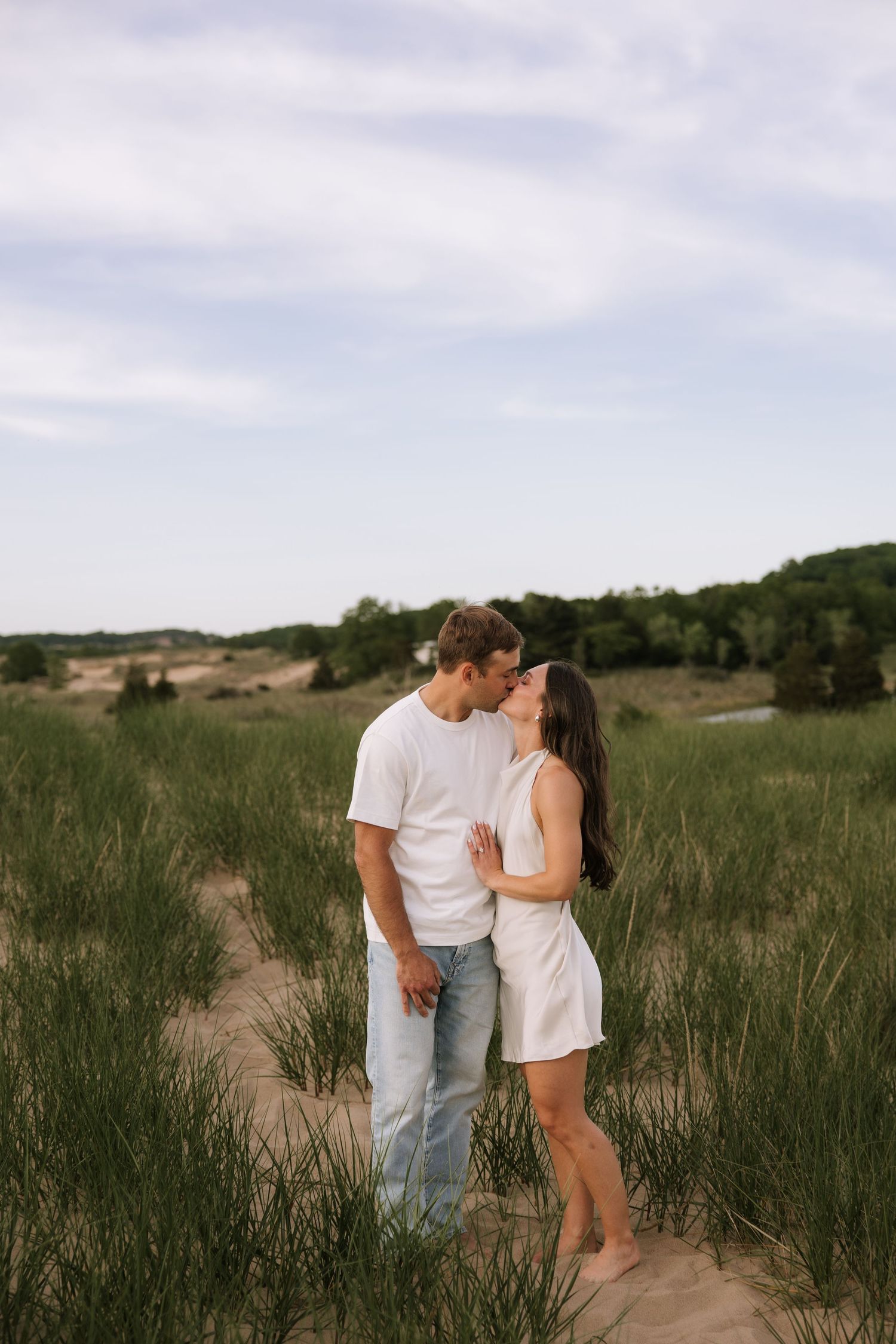 Couple sharing a kiss while standing on sandy dunes surrounded by green grass under a cloudy sky.