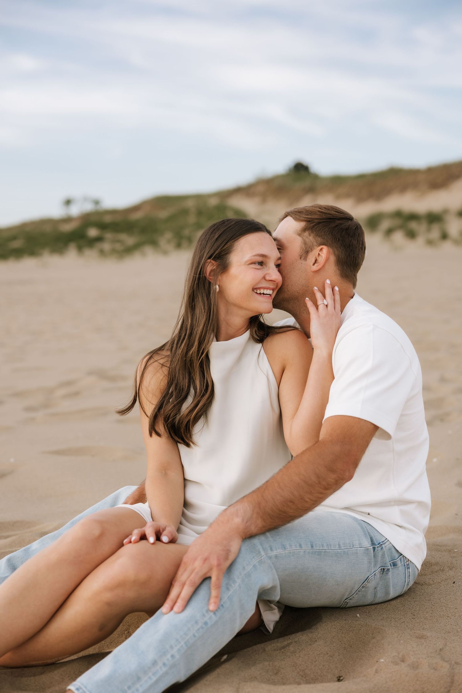 Couple sitting on sandy beach, man whispering to smiling woman with sand dunes in background.