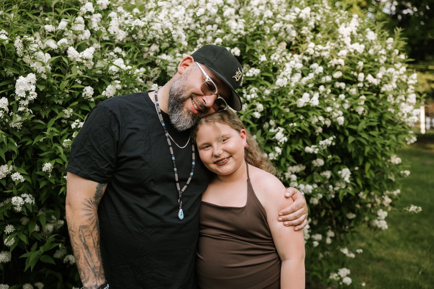 A tattooed man in a black shirt hugs a smiling girl in front of lush white flowering bushes outdoors.