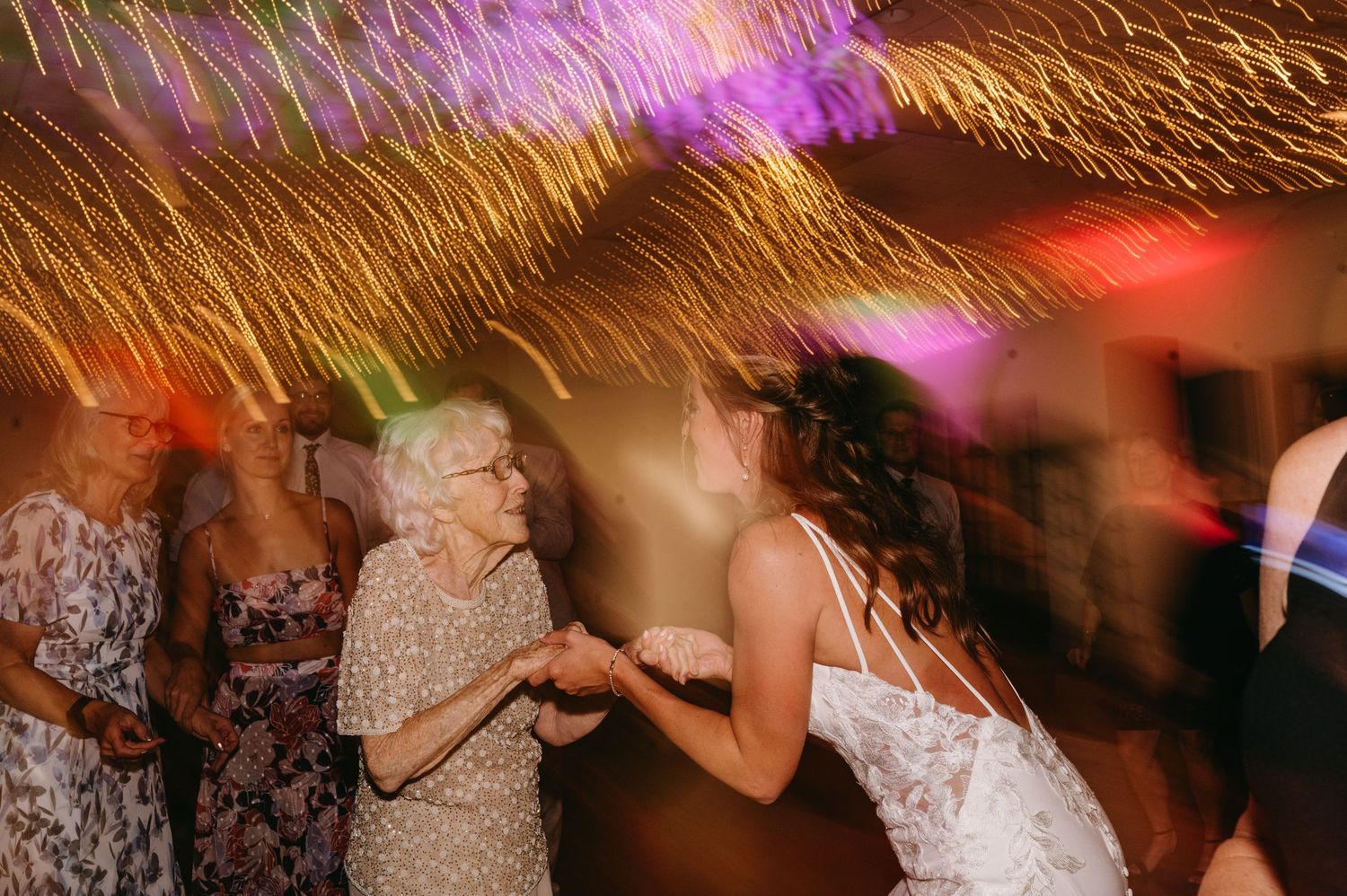 Wedding guests dancing and celebrating on a dimly lit dance floor with motion blur effects and colorful lighting.