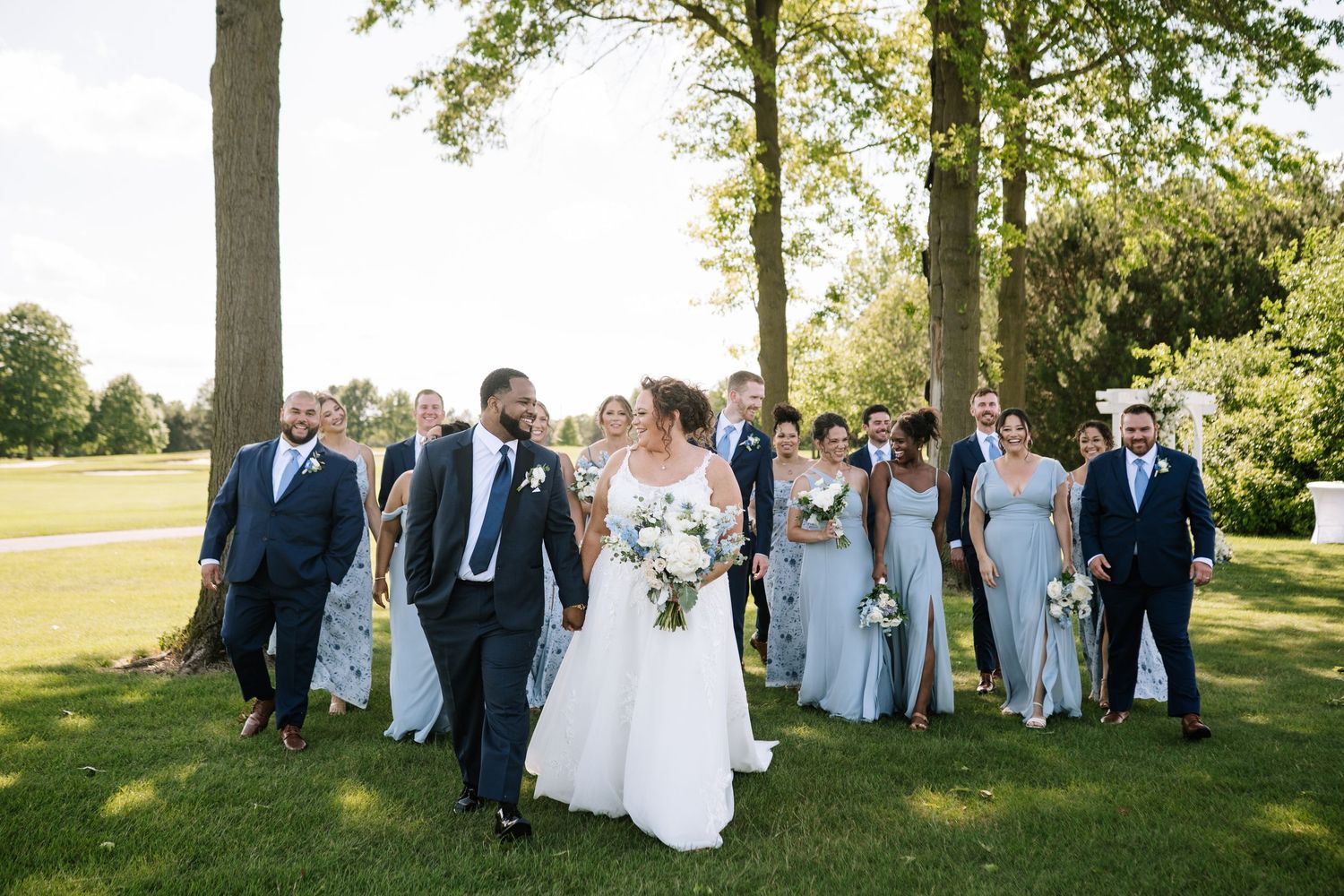 Wedding party gathered outdoors for group photos under tall trees.