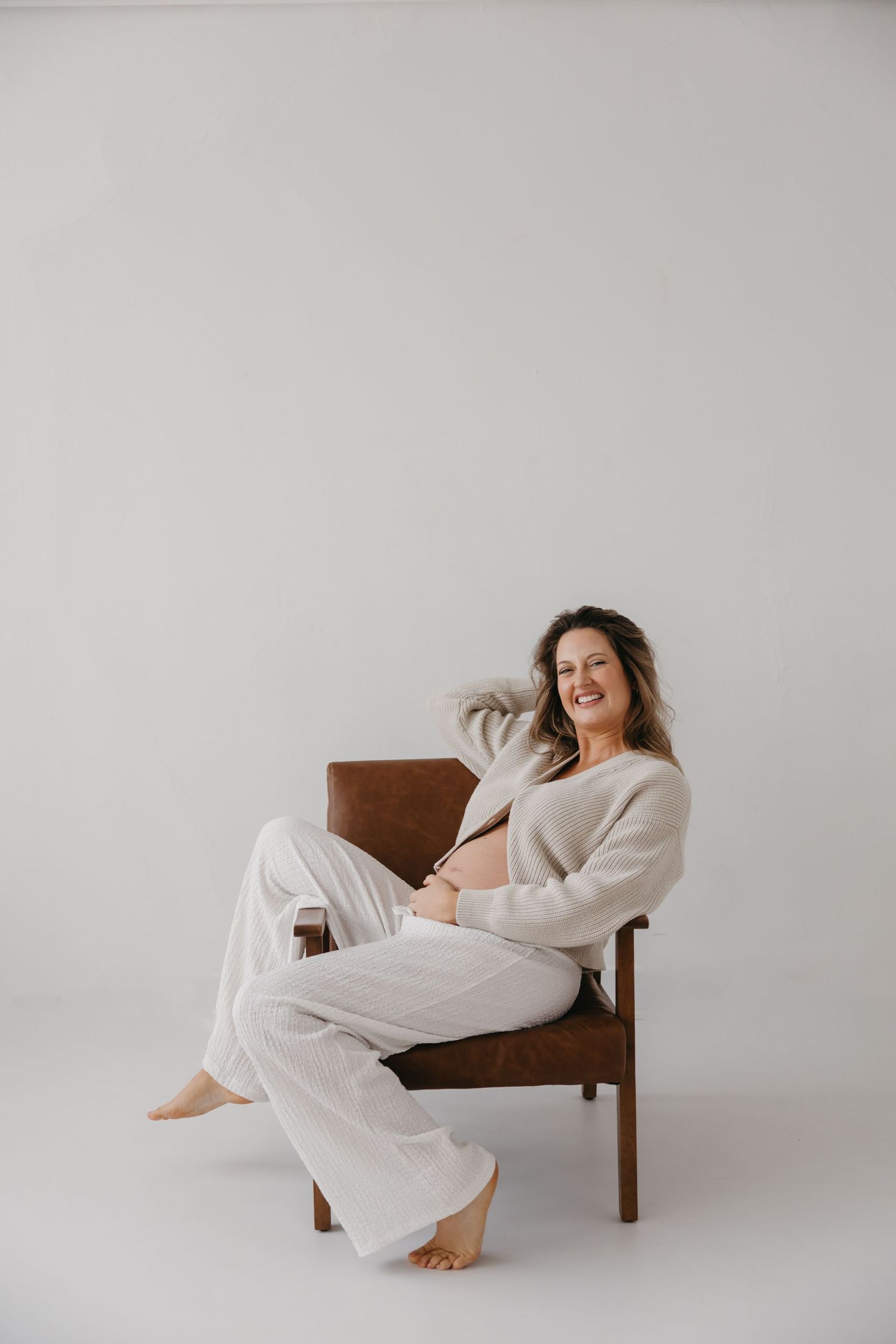 Laughing pregnant woman in cream knit outfit seated in wooden chair against white backdrop.