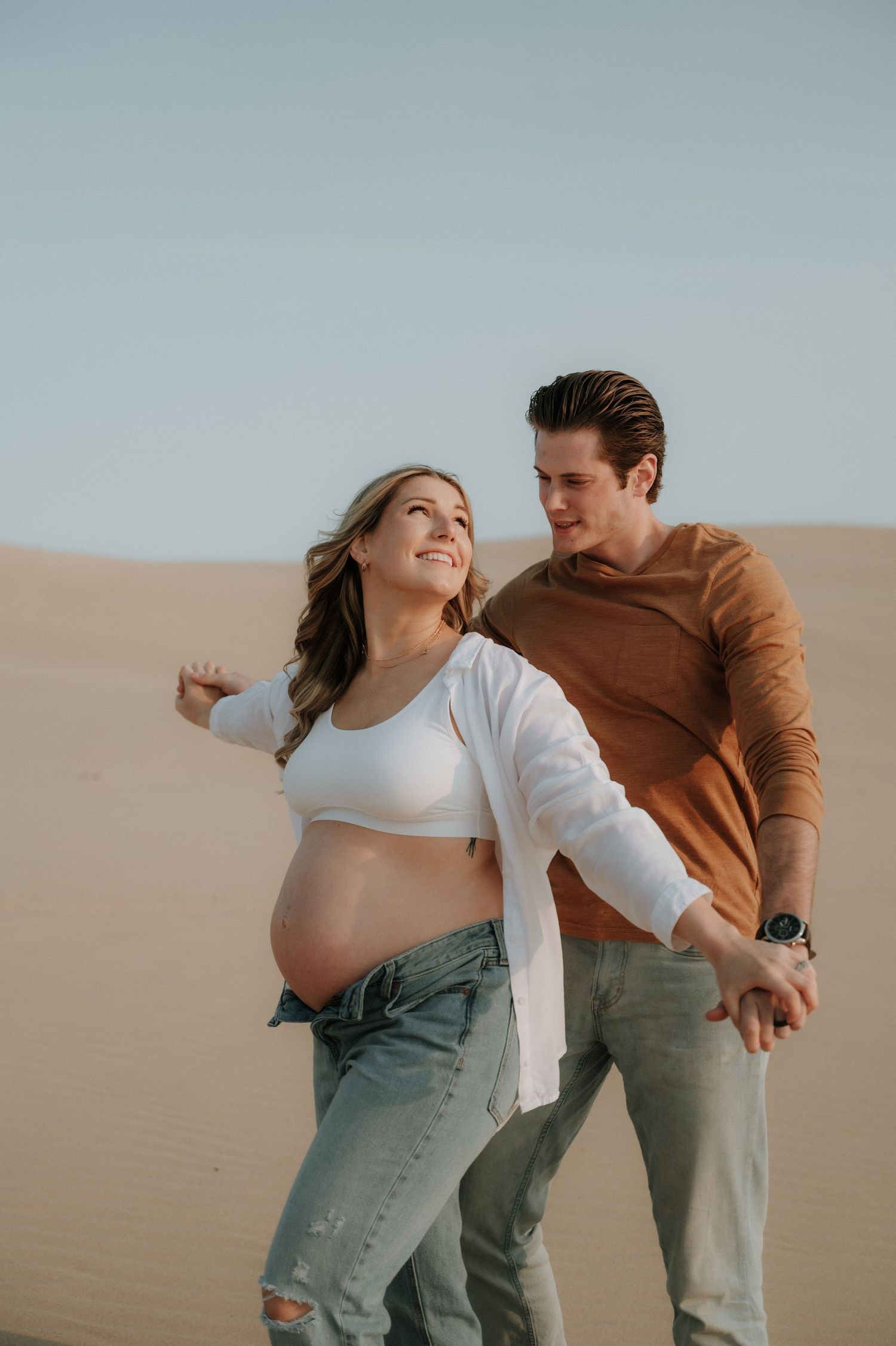 A playful couple laughing together in casual attire against sand dunes at sunset.