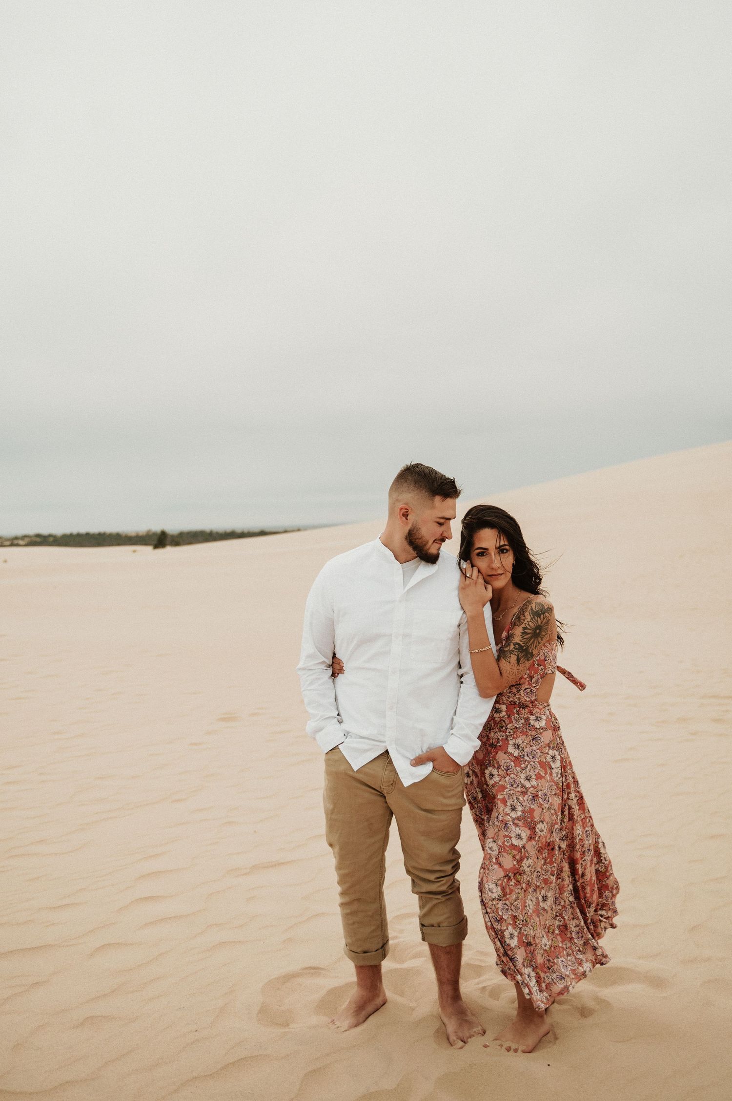 A couple stands close together on sandy dunes wearing coordinated casual outfits.