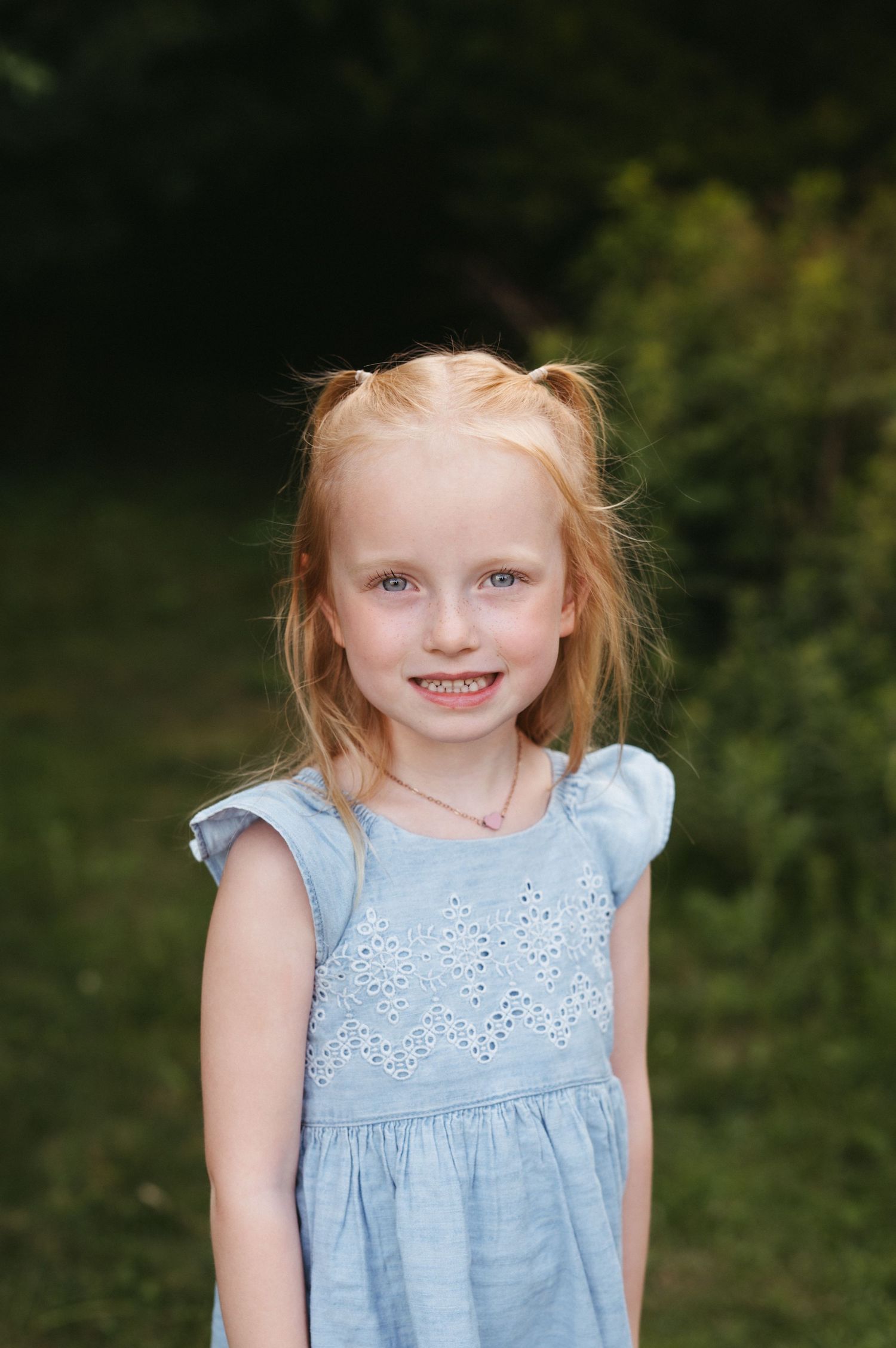A portrait of a young child in a light blue dress standing in front of green foliage.