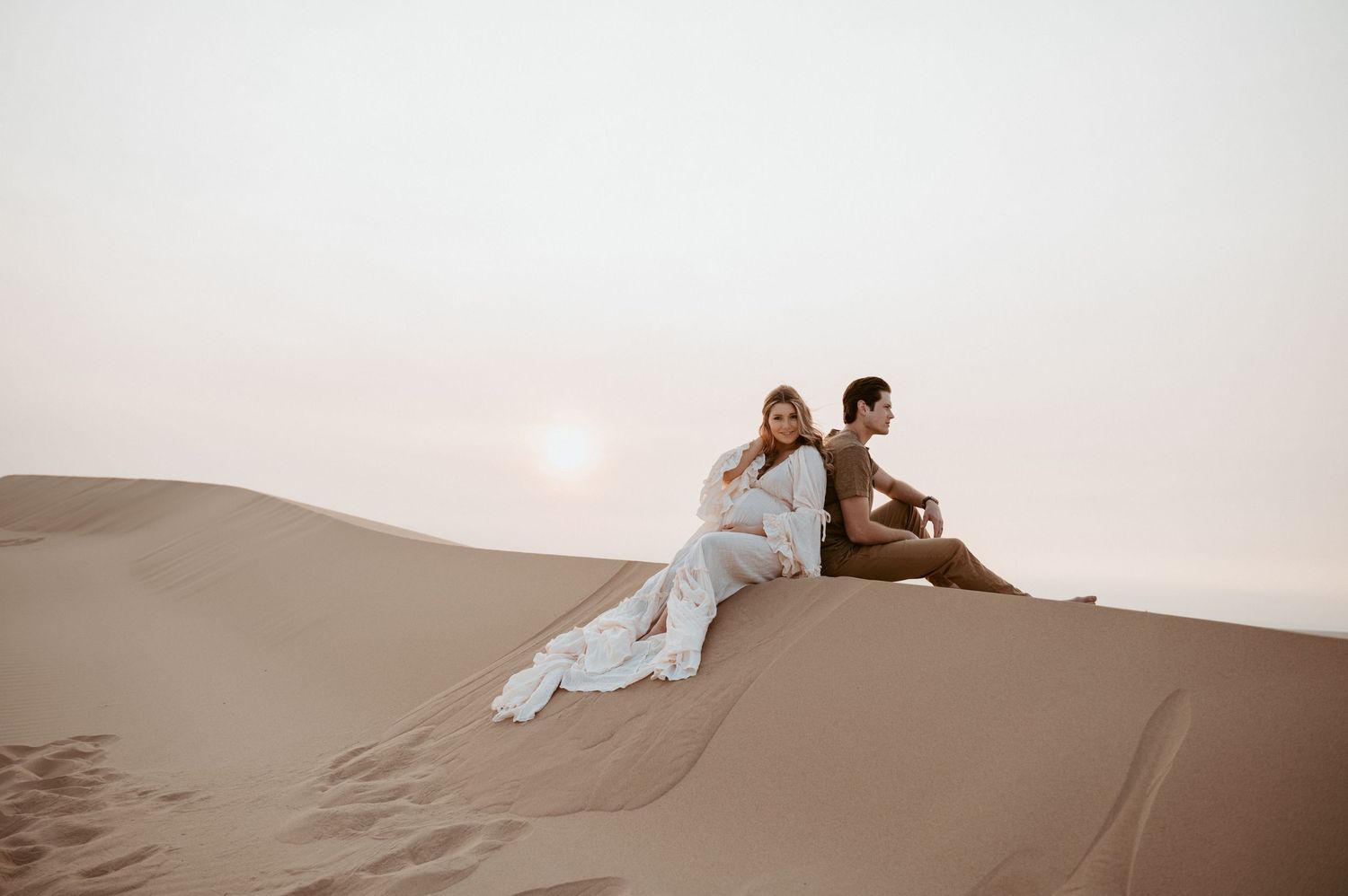 Two people share an intimate moment while sitting on desert sand dunes at sunset.