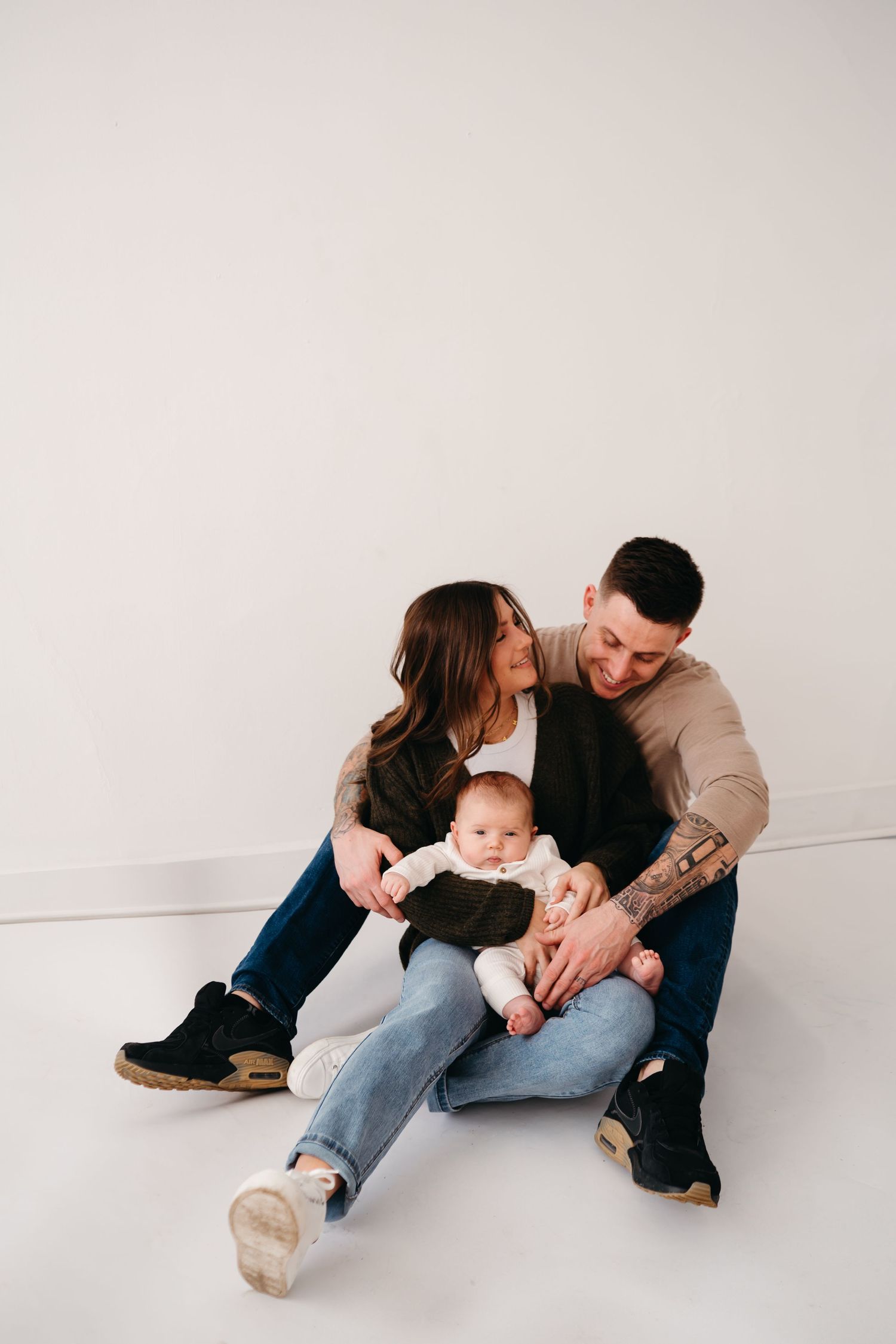 Family of three sits on white studio floor, sharing affectionate moment while holding their infant child.