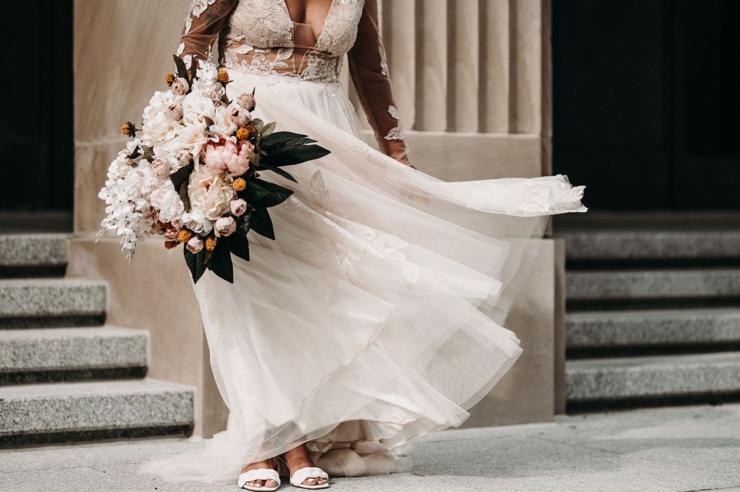 The hem of an elegant white wedding gown swirls with movement beside a delicate pink and white bouquet.