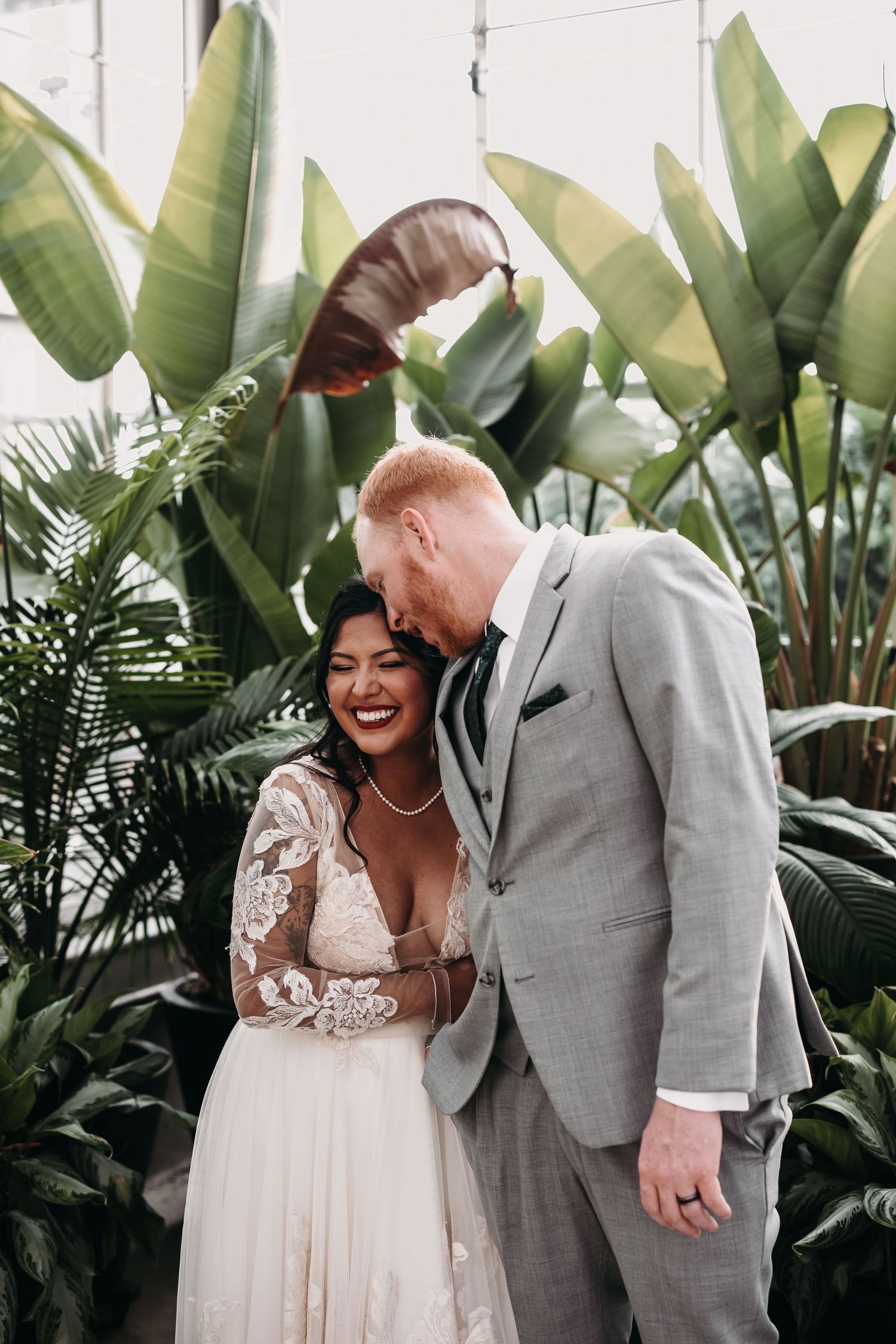 Newlyweds share an intimate moment among tropical plants in a greenhouse setting.