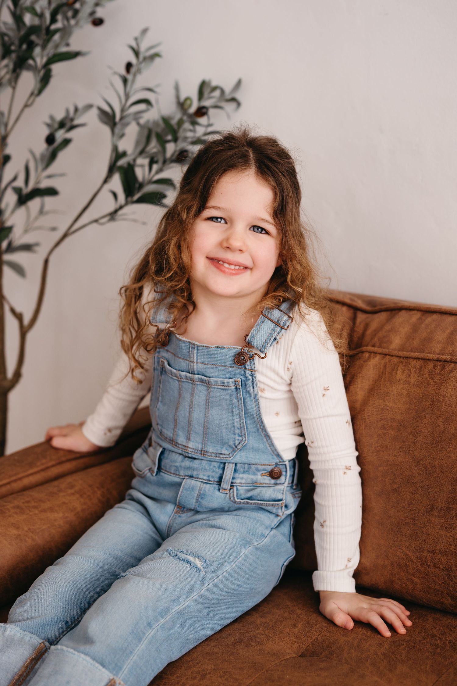 Child wearing blue denim overalls and white shirt sits smiling on brown leather couch with plant in background.