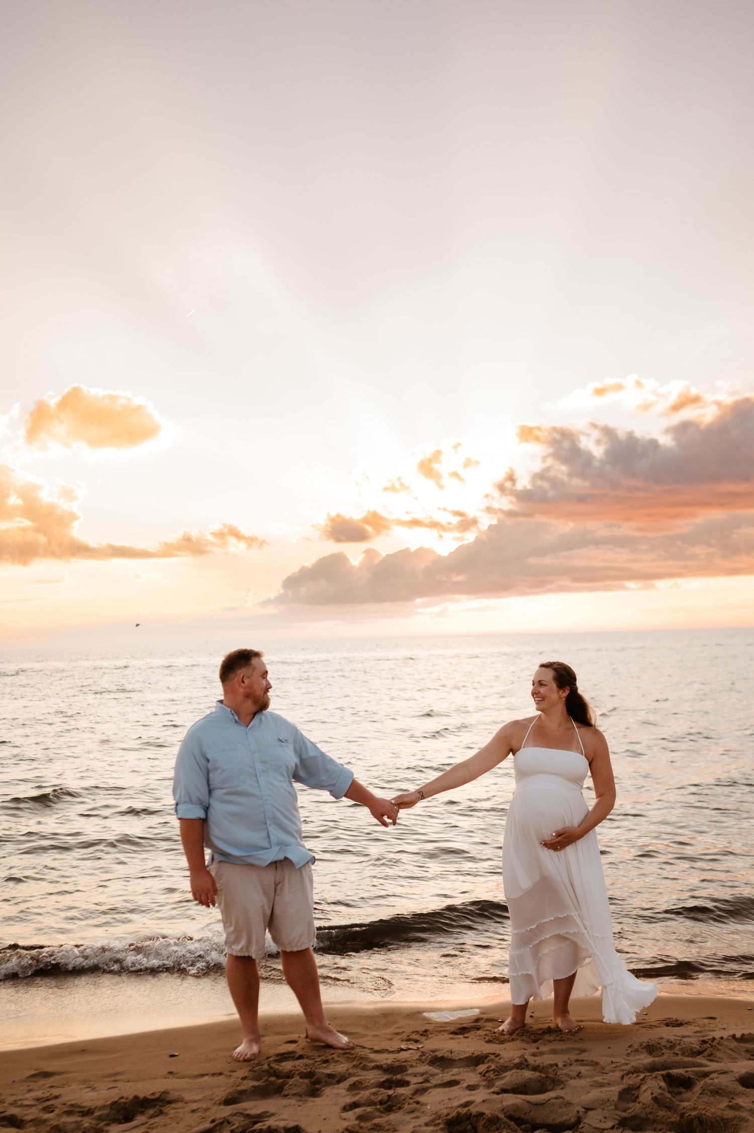 A couple holds hands on a sandy beach at sunset with orange clouds and ocean waves in the background.