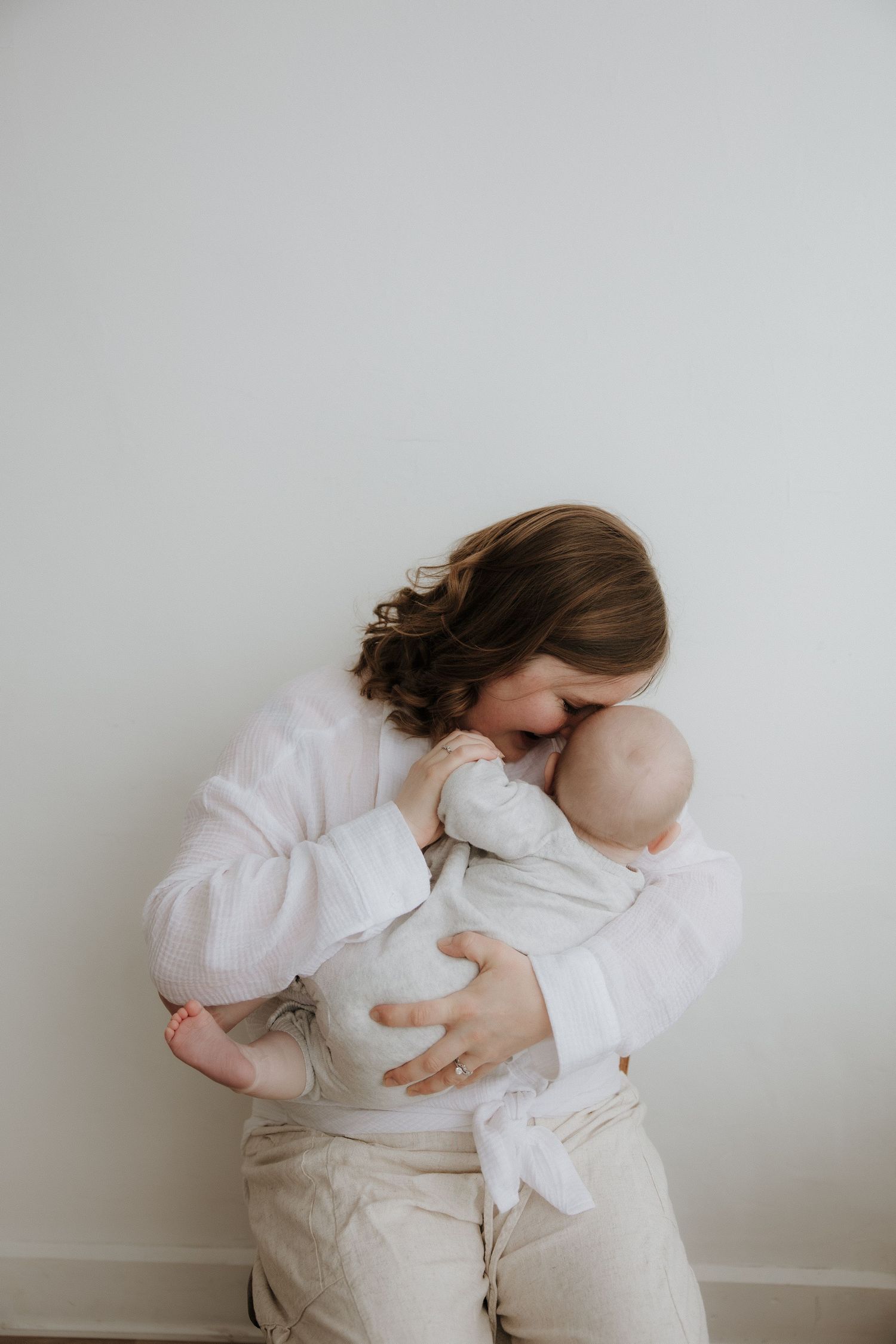 A mother in white linen gently nuzzles her newborn baby, sharing a tender moment against a soft white background.