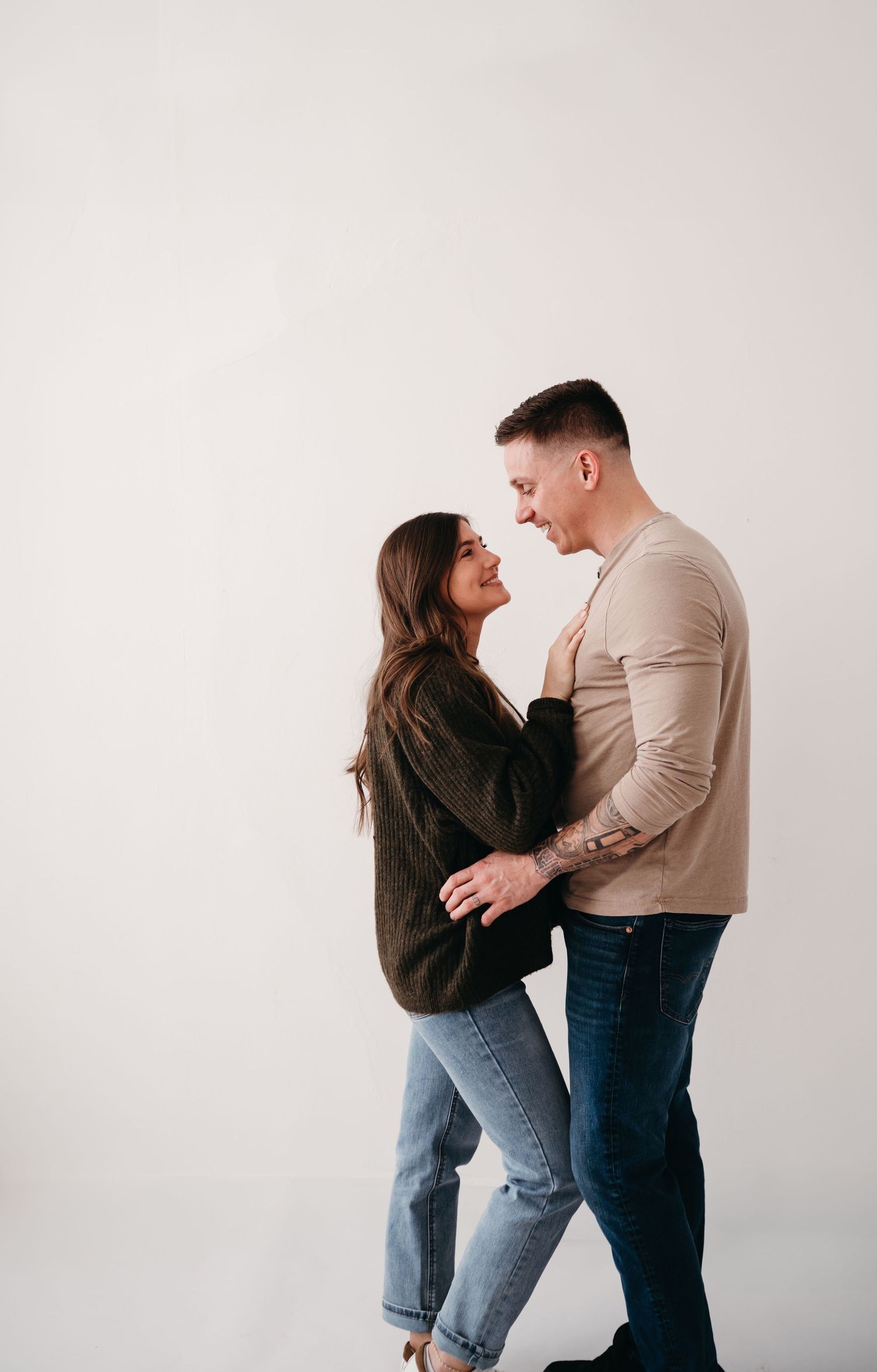 A couple shares an intimate moment while standing against a white background, dressed casually in jeans and neutral tones.