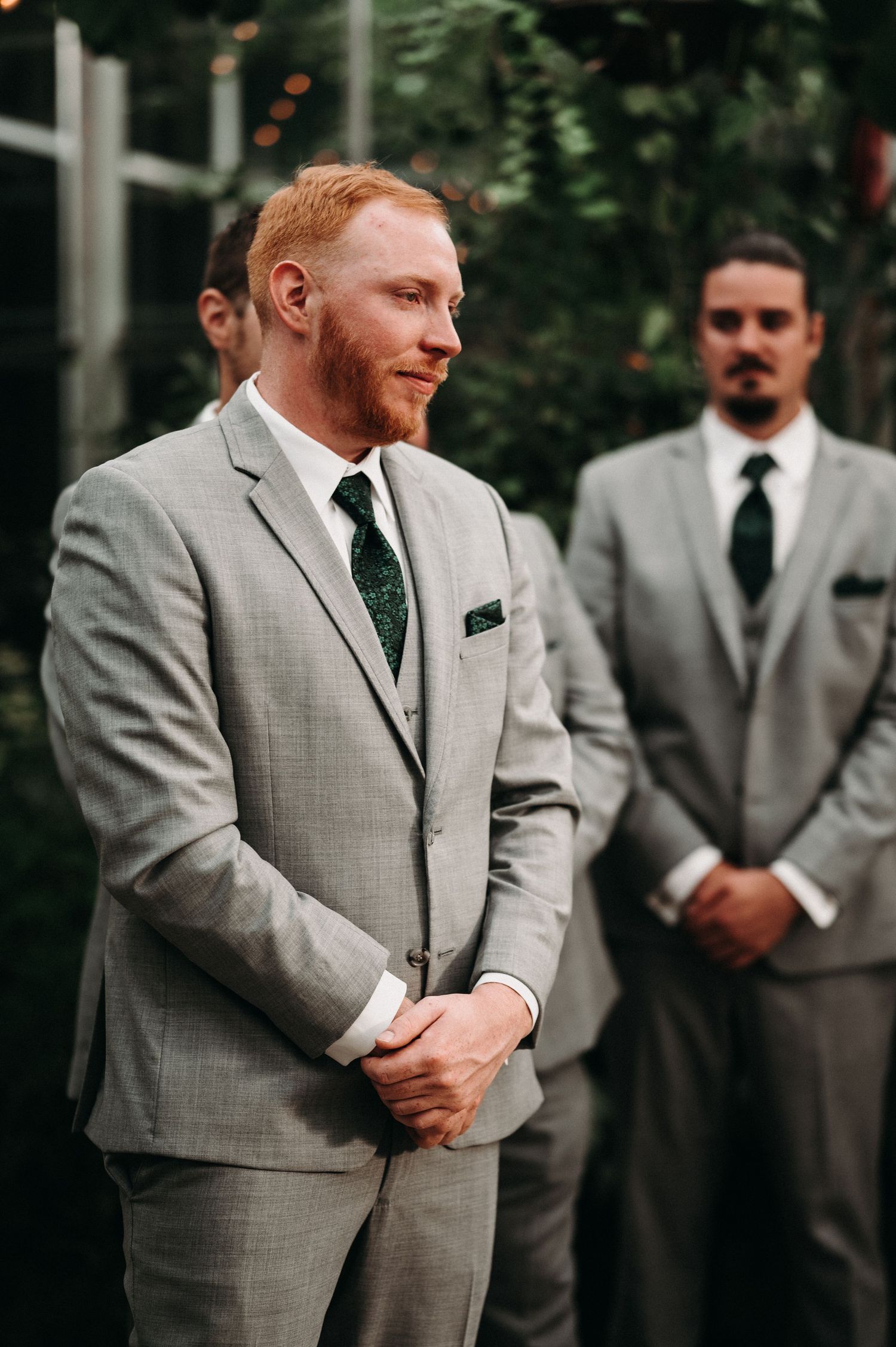 Groomsmen in gray suits standing during outdoor wedding ceremony.