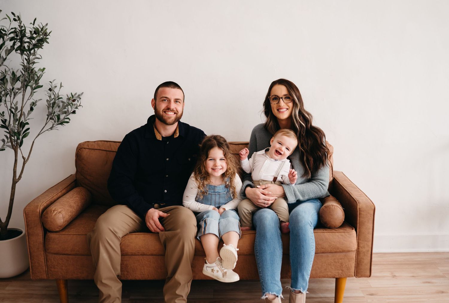 A family sits together on a brown leather couch in a minimalist living room with white walls and potted plants.