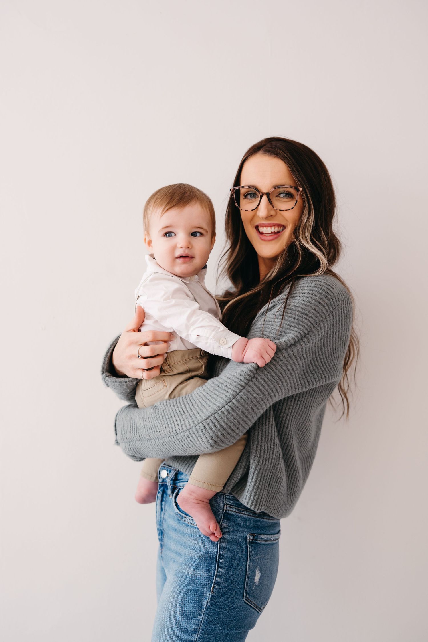 A parent wearing a grey sweater and jeans holds a baby in white against a light background in a heartwarming pose.