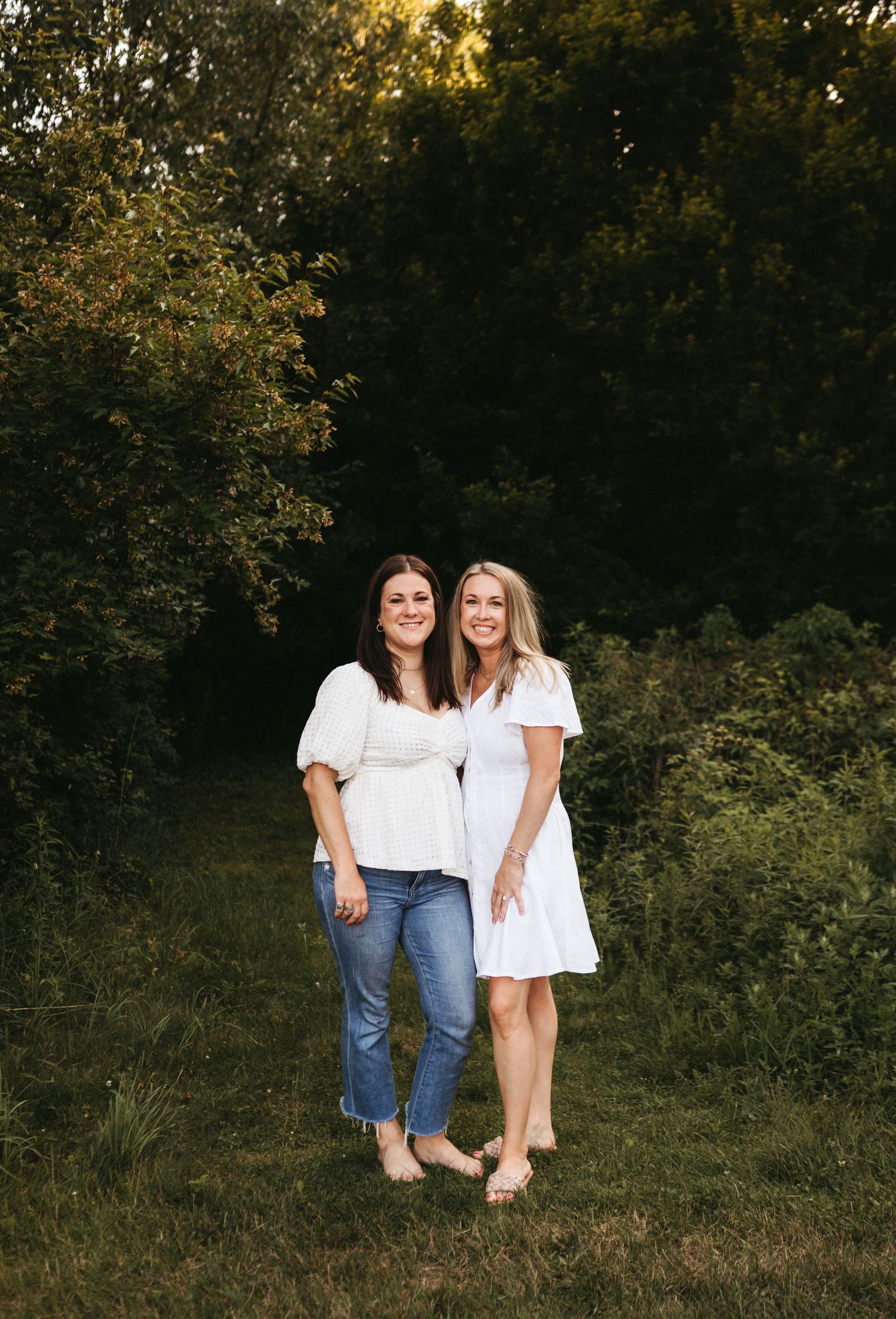 Two friends pose together at sunset in a natural outdoor setting wearing white tops and summer attire.