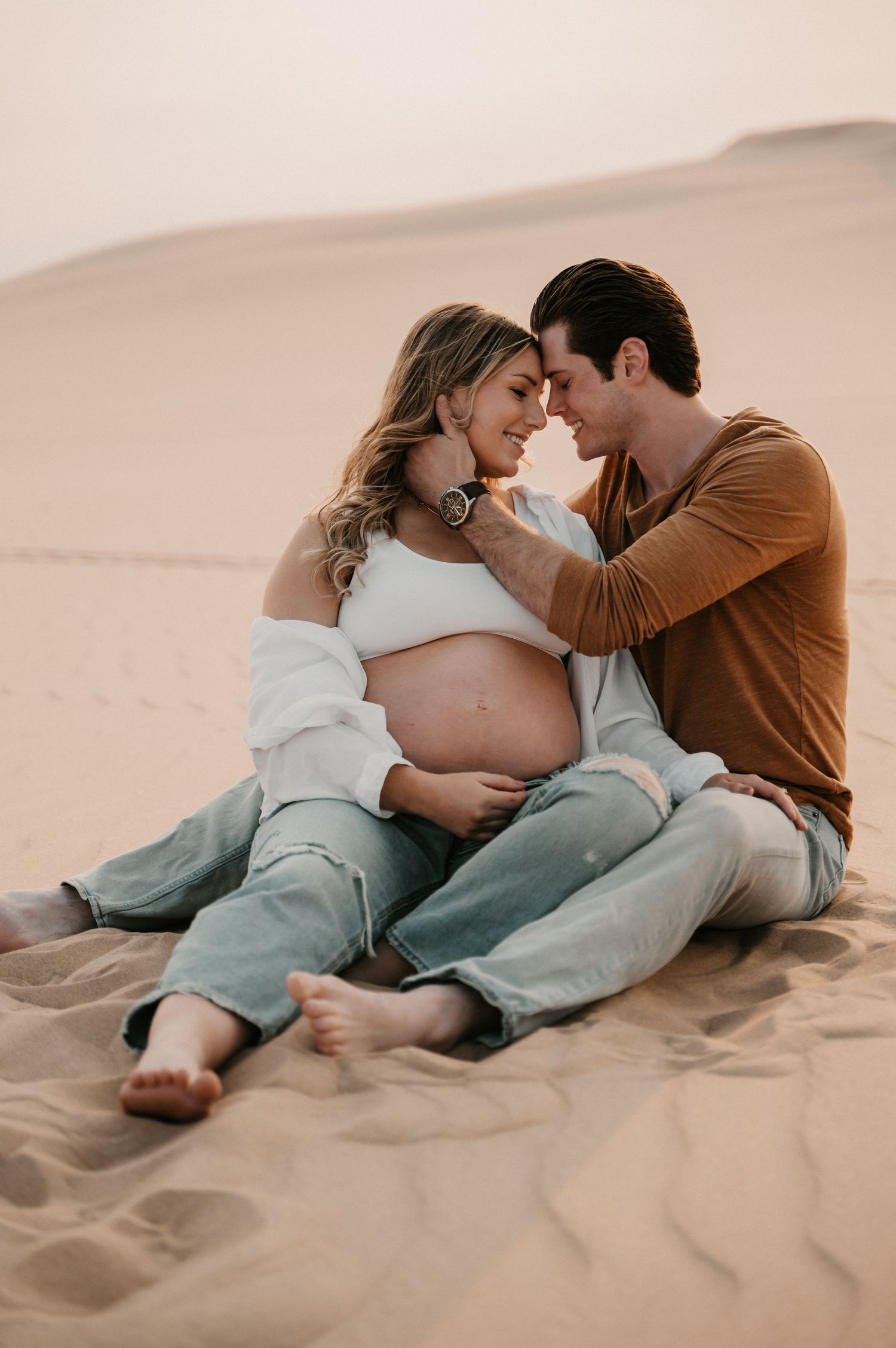 Expecting couple shares tender moment while sitting on desert sand during sunset photoshoot.