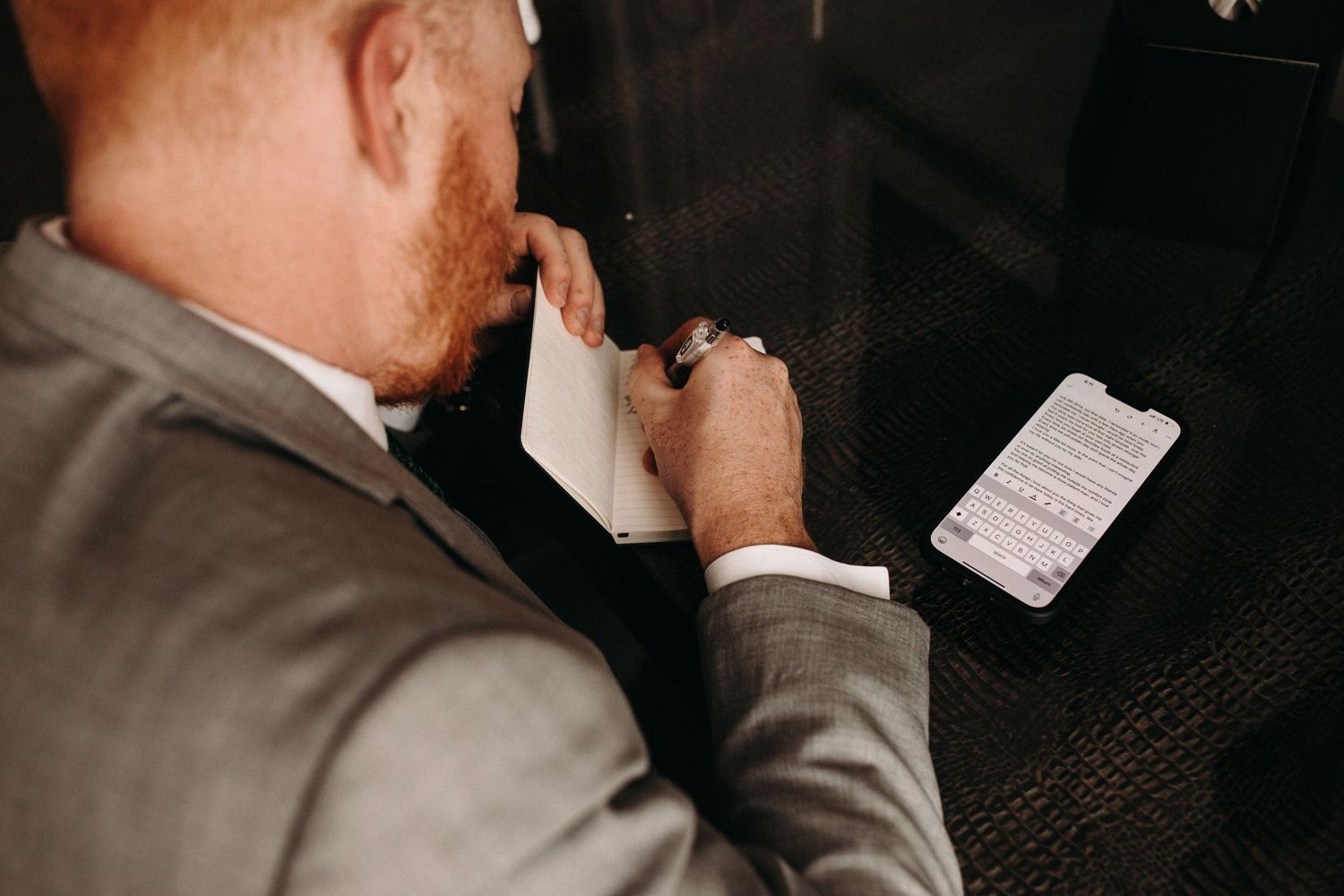 Person in grey suit writing notes at a dark wooden table.