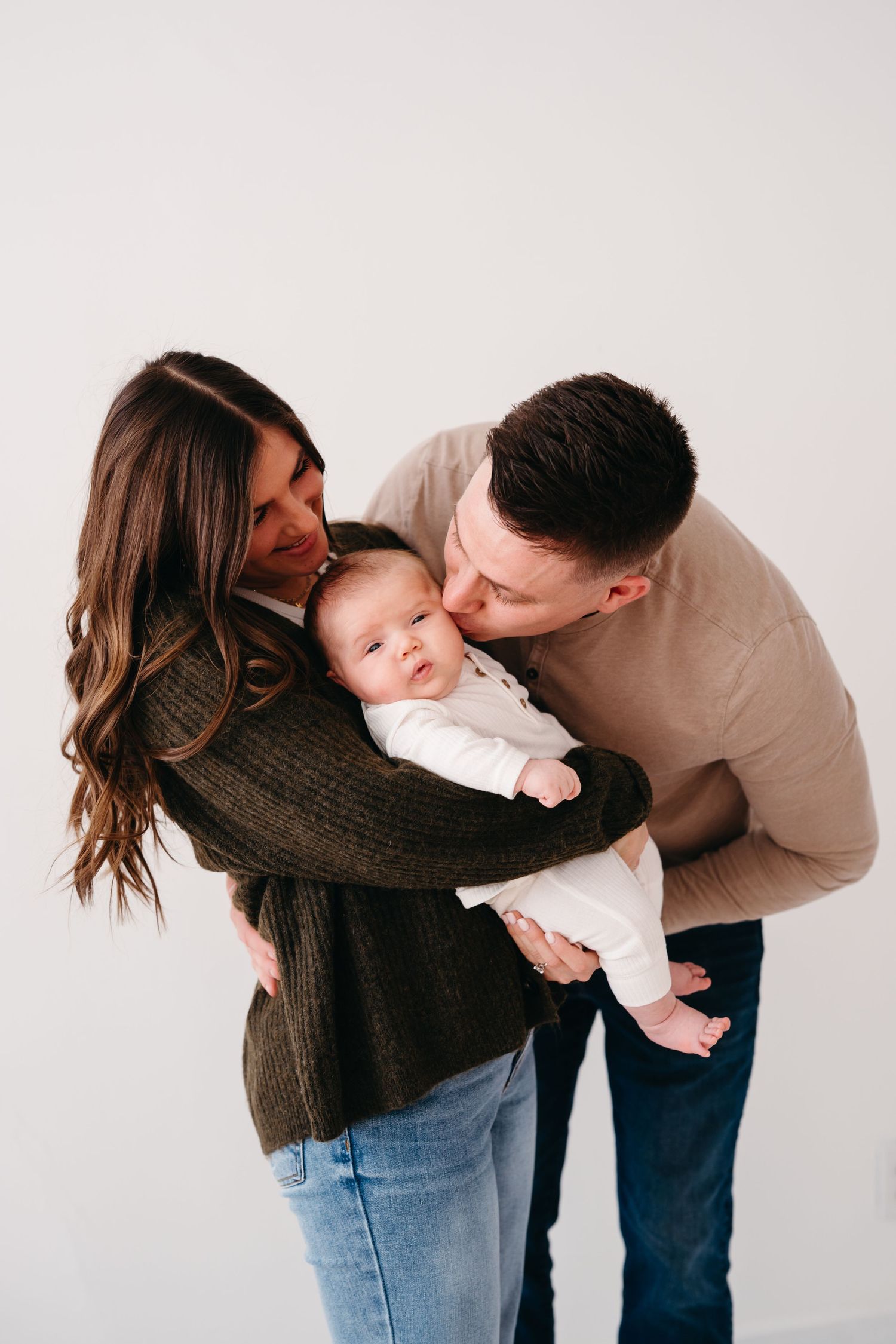 Parents gazing lovingly at baby in white onesie against neutral background.