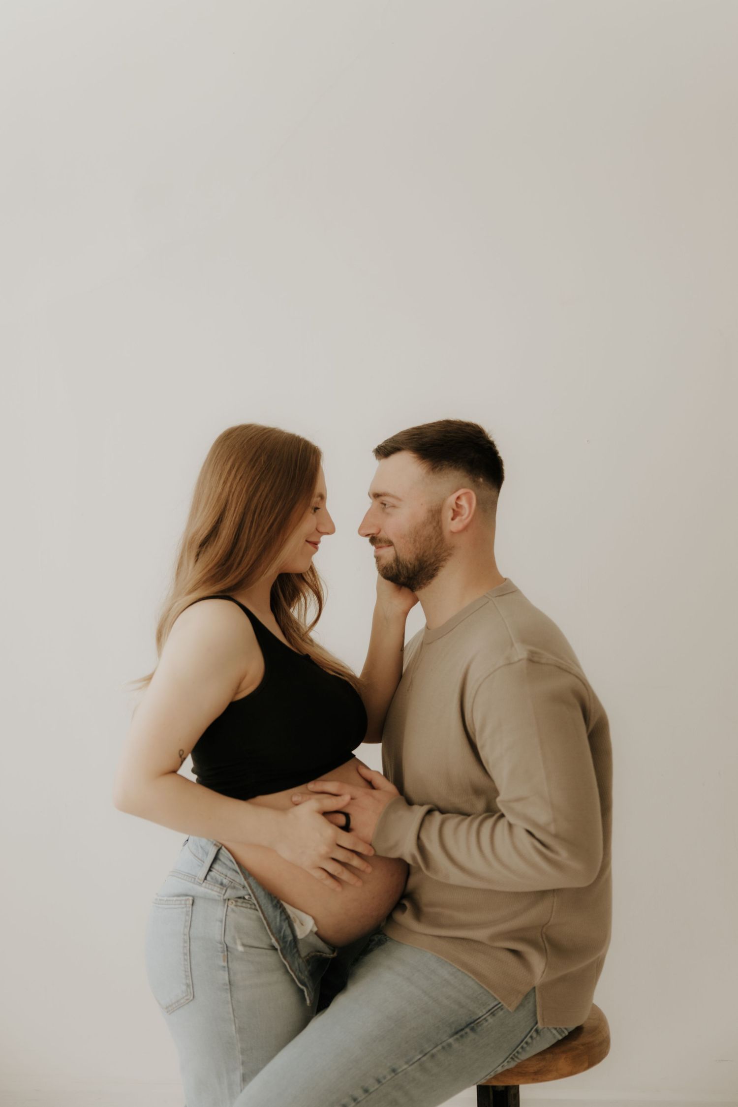 Intimate maternity moment captured against white wall in casual attire.