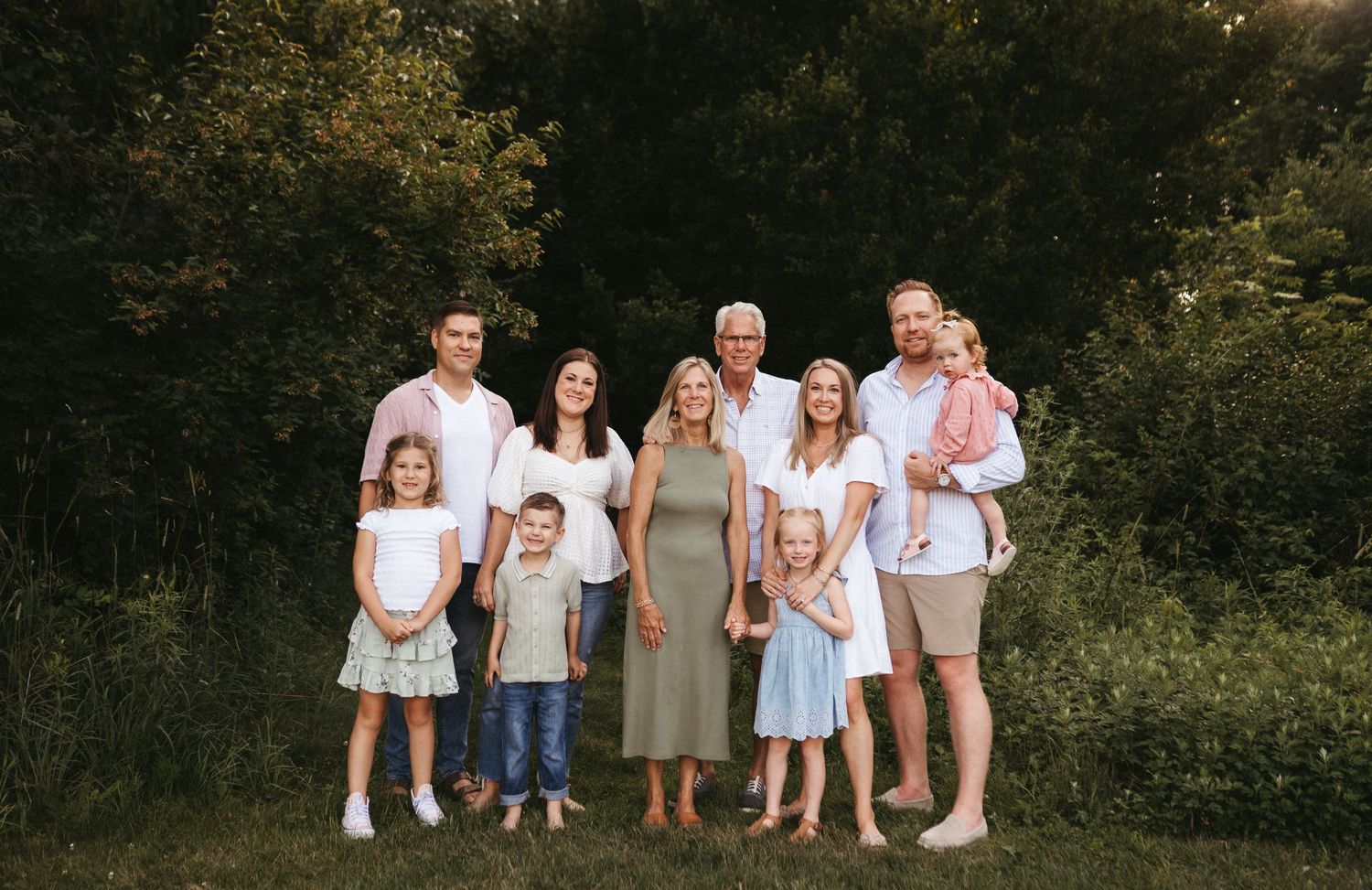 Extended family group poses together for a portrait outdoors against a wooded backdrop.
