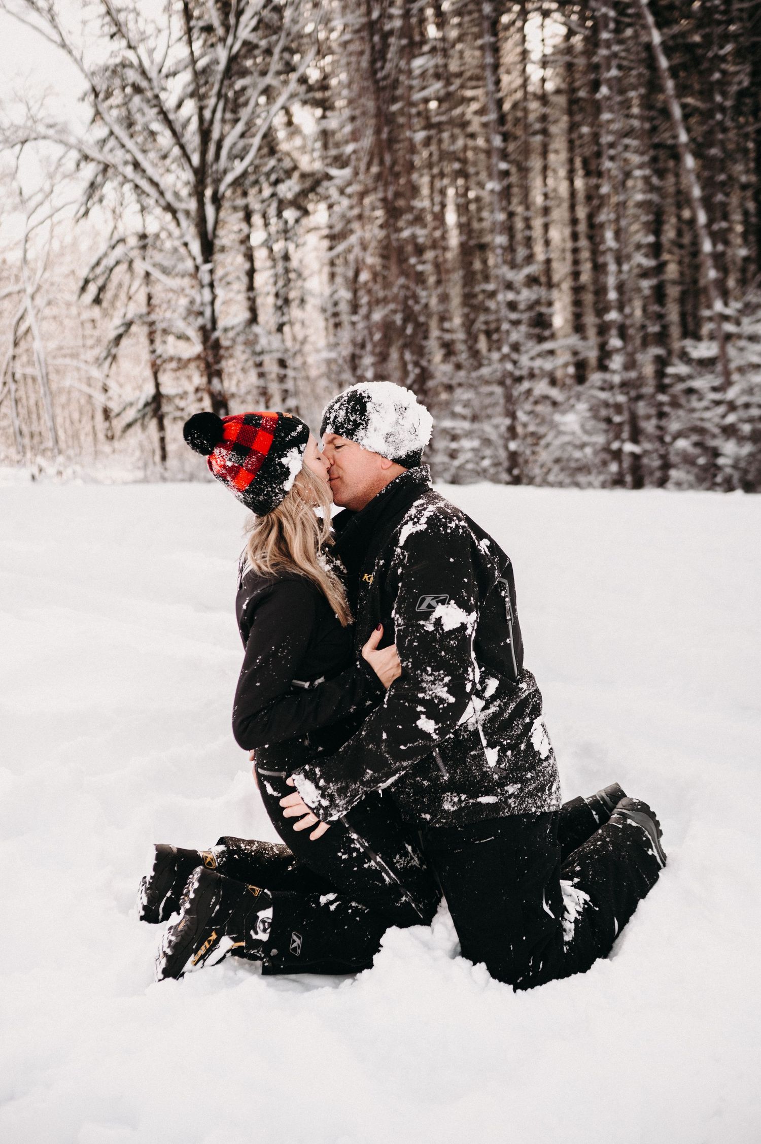 A winter engagement moment captured in a snowy forest with matching buffalo plaid beanies.