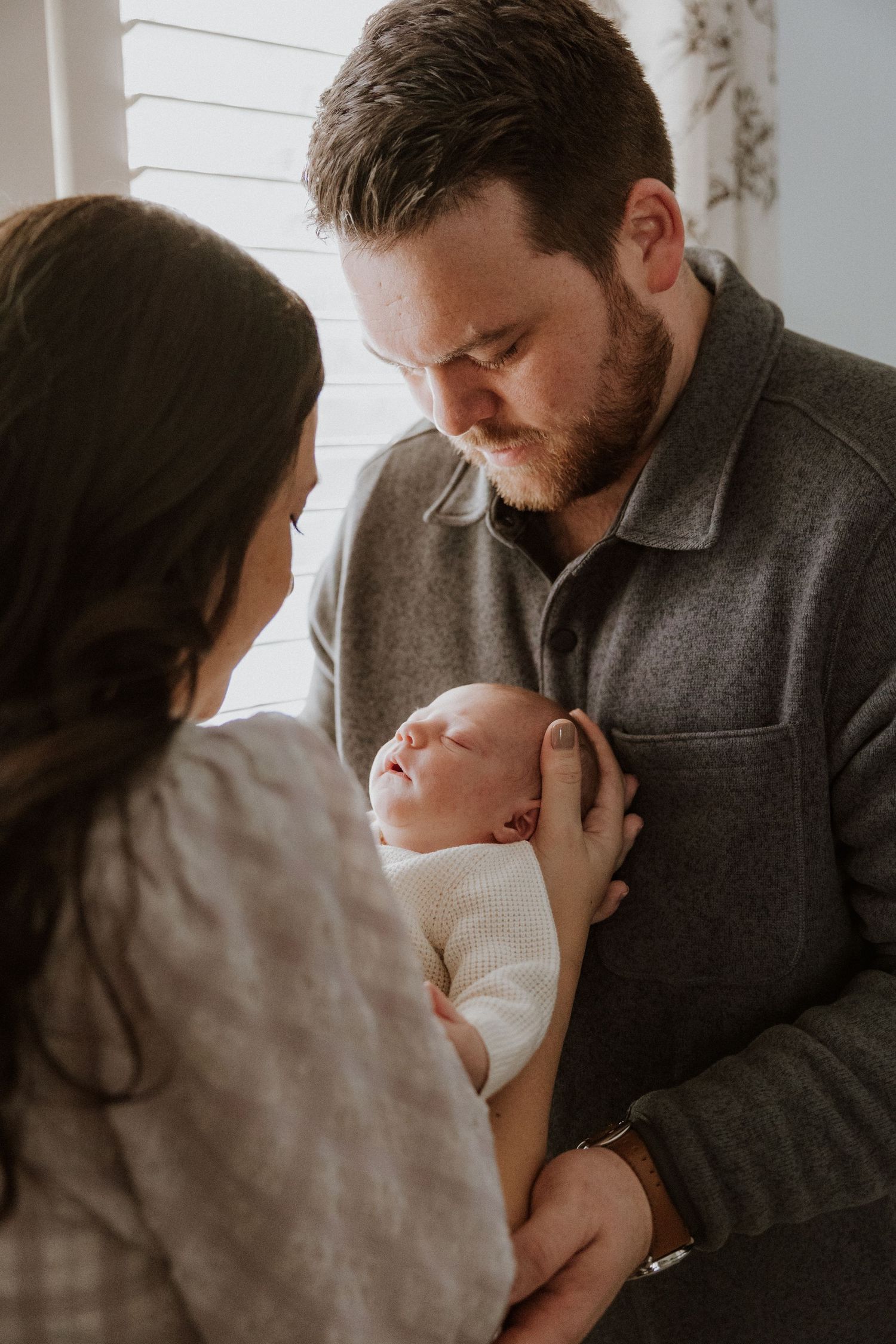 Intimate family moment with baby in white clothing by a bright window.