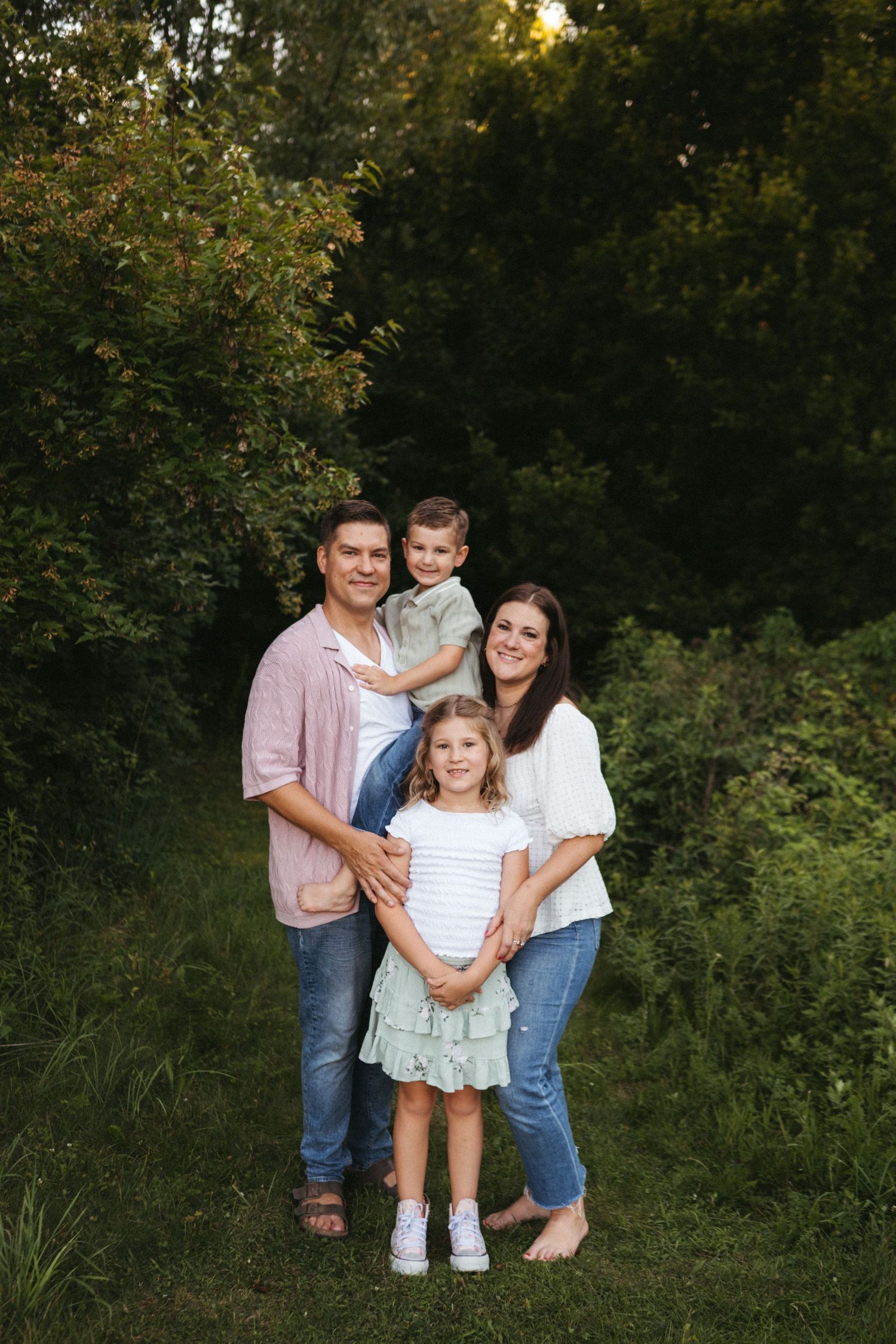 A family of four poses together outdoors in coordinated casual outfits against trees at sunset.