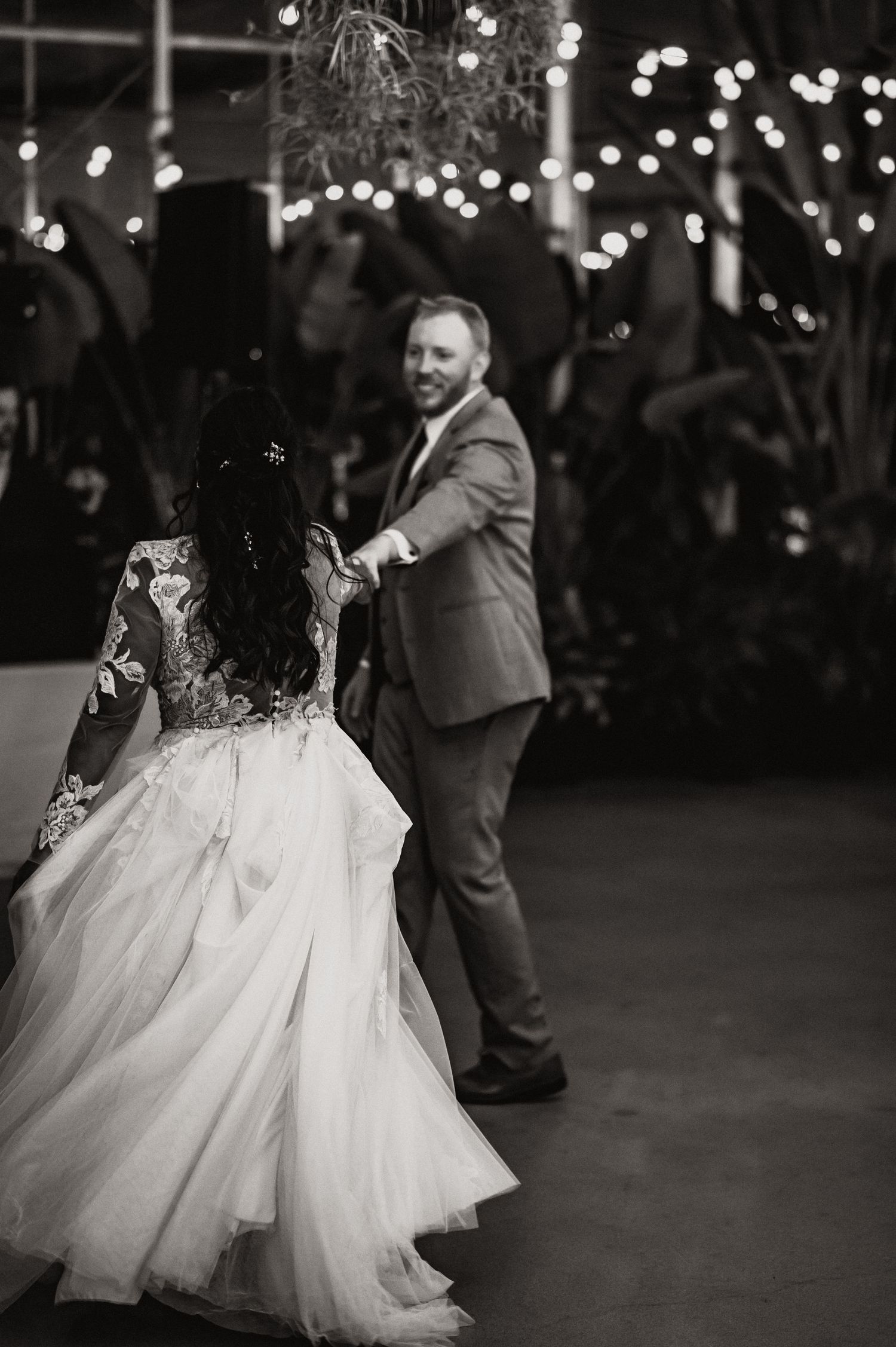 Black and white photo of couple dancing at wedding reception with string lights in background.