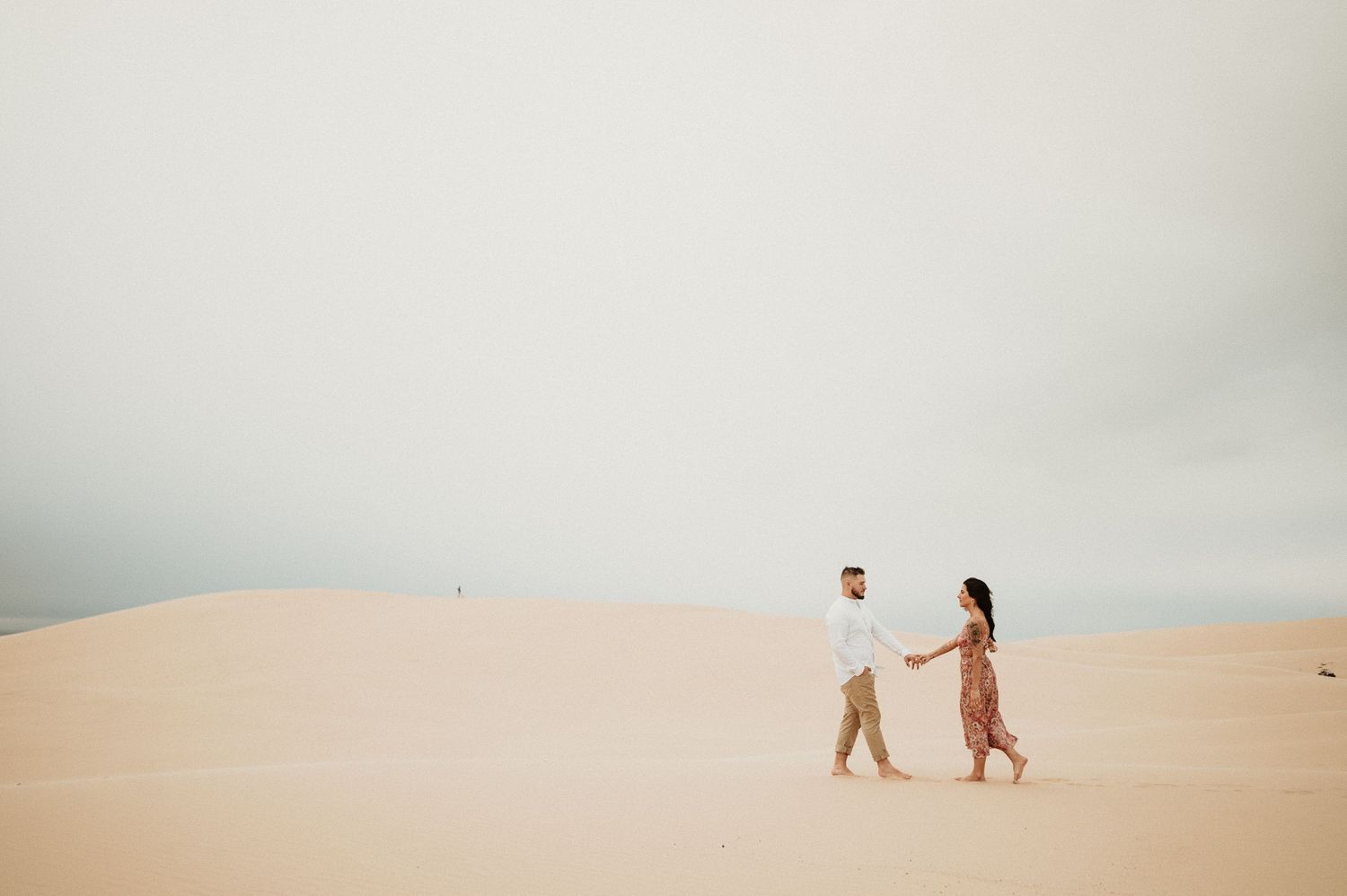 A distant view of two people walking hand in hand across vast desert sand dunes under cloudy skies.