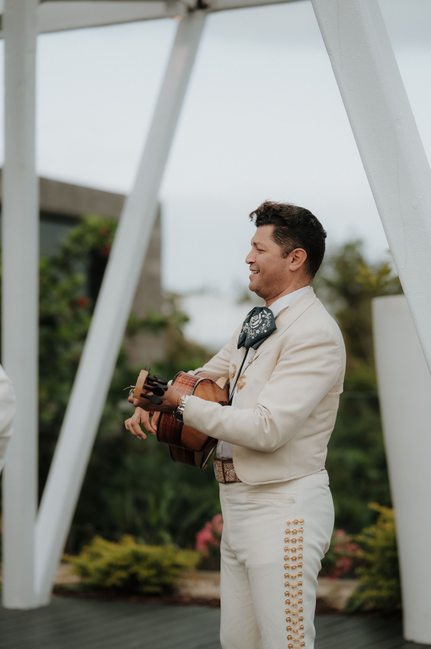 Mariachi musician in white traditional suit plays guitar against white architectural backdrop.