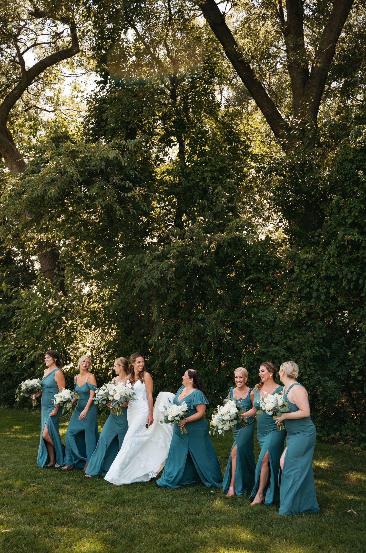 Wedding party members in teal blue dresses sit in a row on grass beneath towering trees.