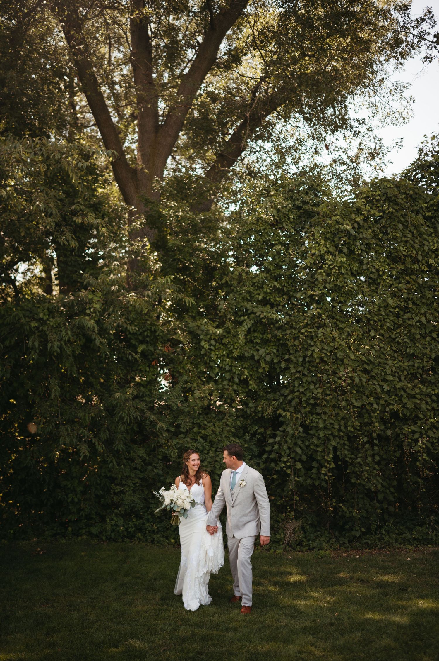 A couple in wedding attire walking through a lush garden setting with trees and greenery in the background.
