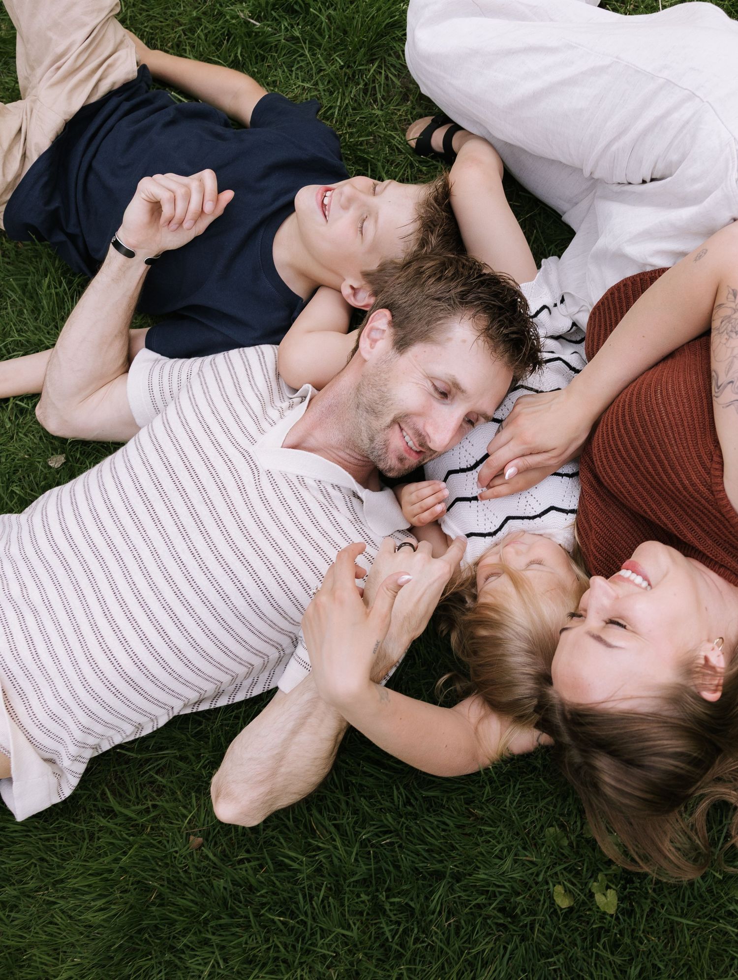 Happy family of four lying on green grass, laughing and bonding together in a joyful outdoor moment.