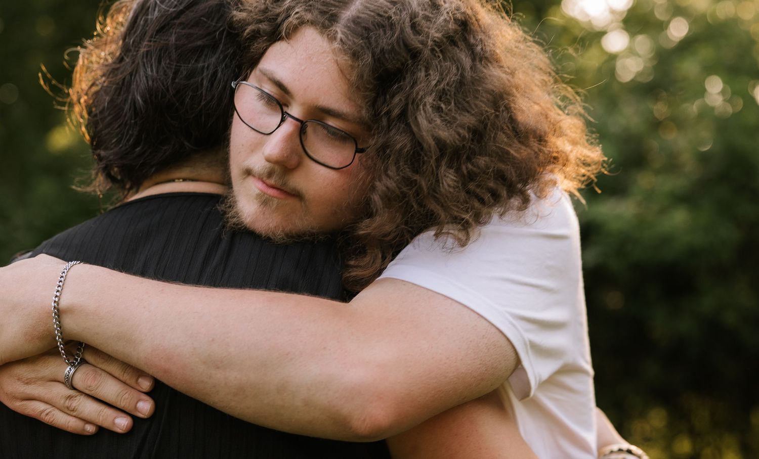 Two young people sharing a warm embrace outdoors, one wearing glasses and a white shirt.