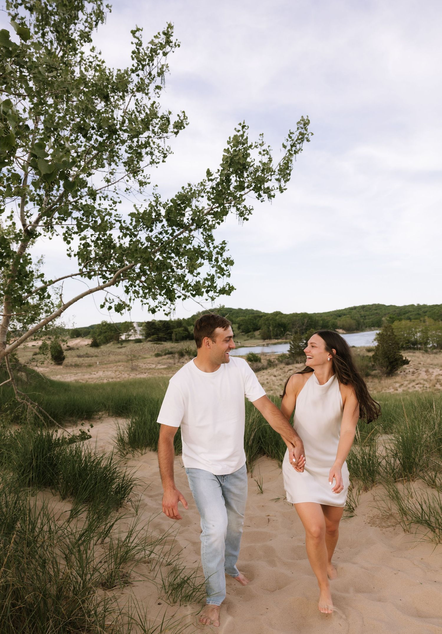 Smiling couple holding hands and walking barefoot on sandy beach dunes near a tree and lake.