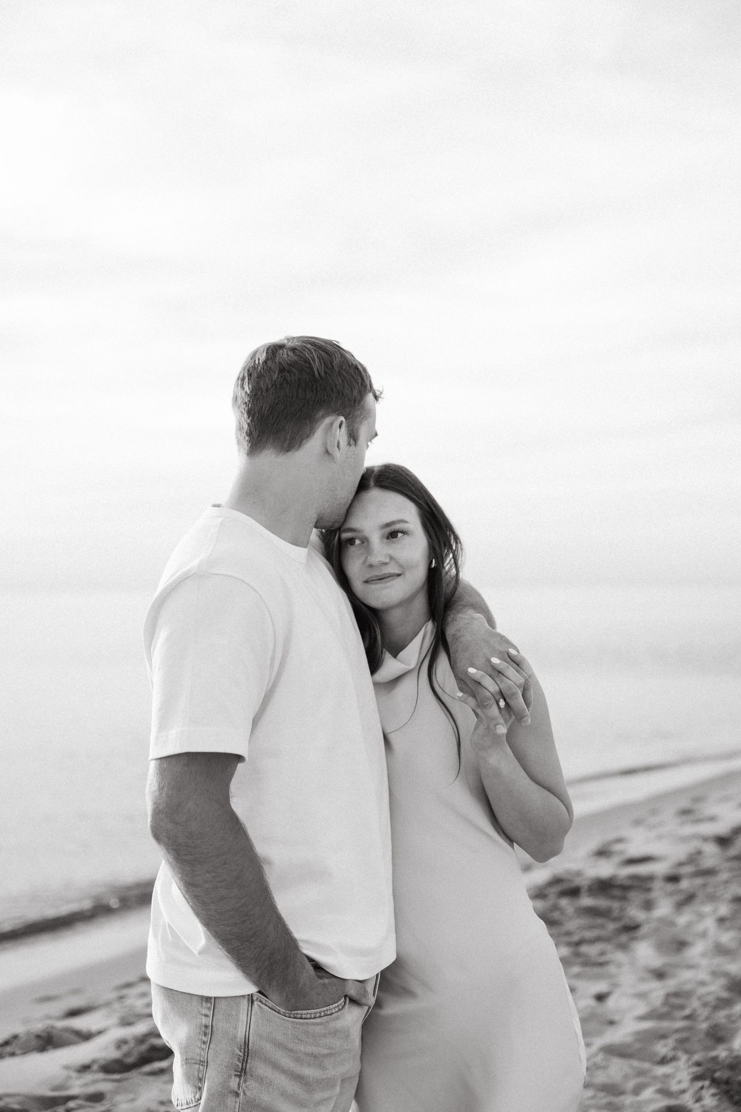 Black and white romantic beach photo of couple embracing, woman gazing up at man near the shoreline.