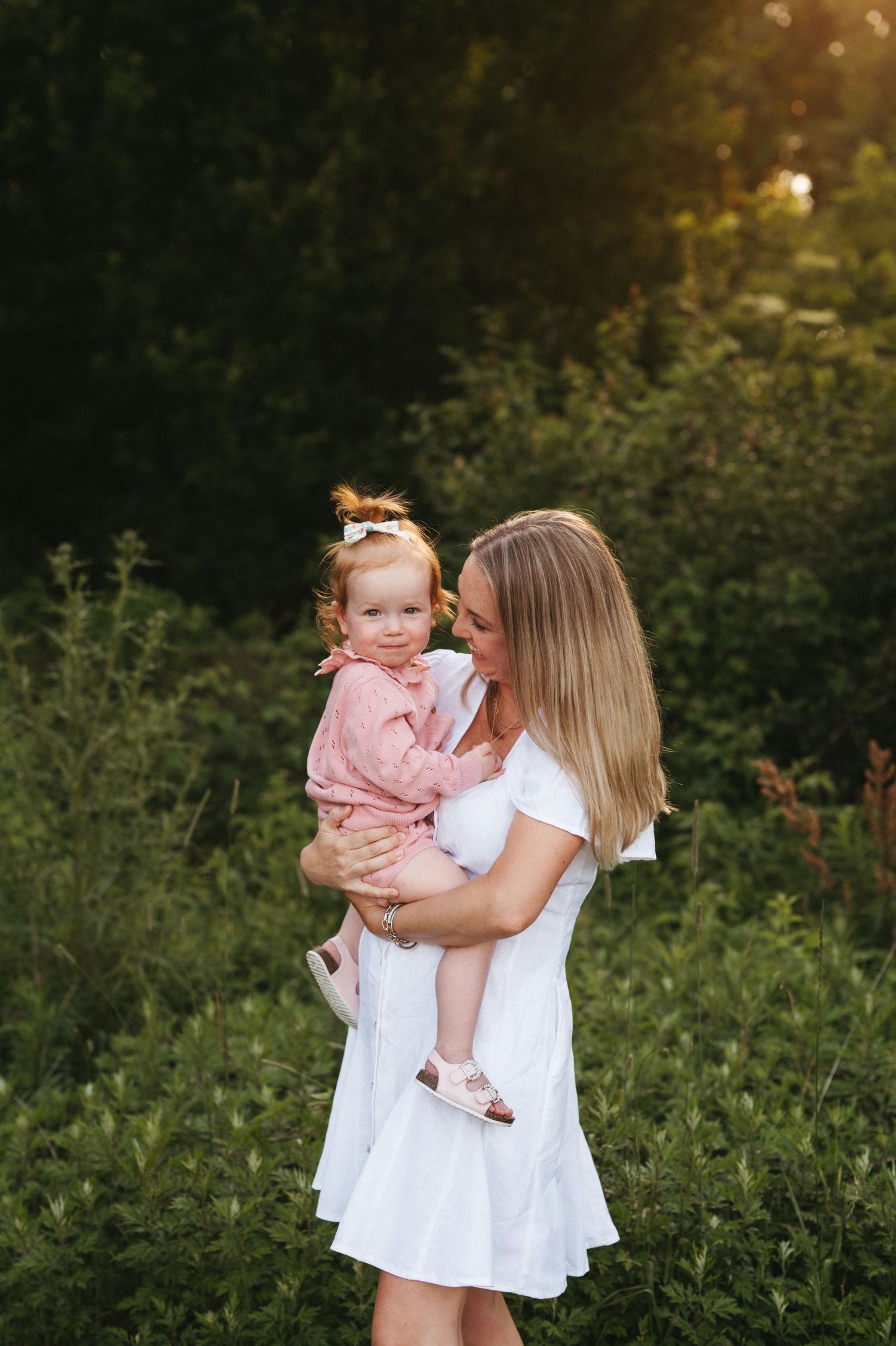 Woman in white dress holding young child in pink outfit outdoors against lush natural backdrop at sunset.