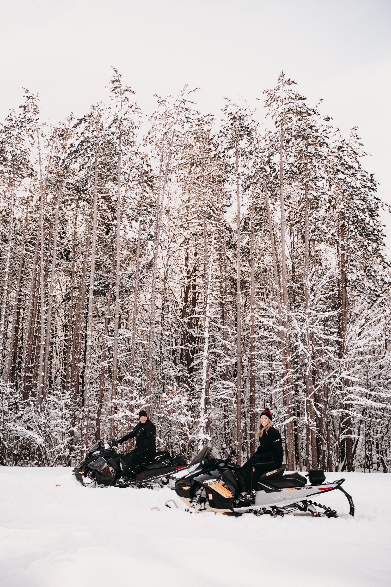 Two snowmobiles parked in a snow-covered forest with tall snow-laden trees.