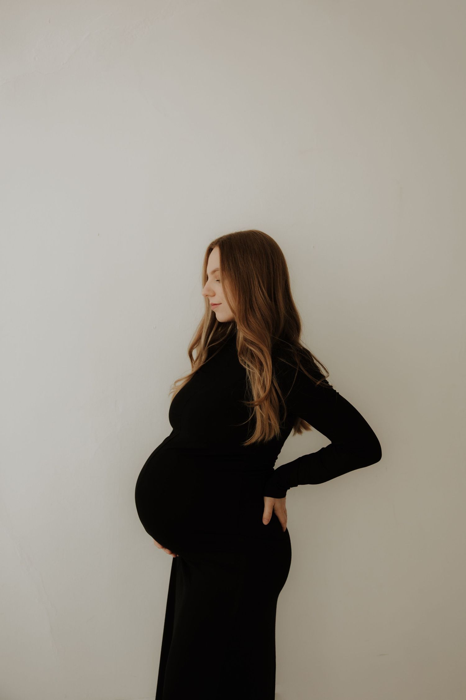 Artistic maternity portraits in black dress with dramatic window lighting.