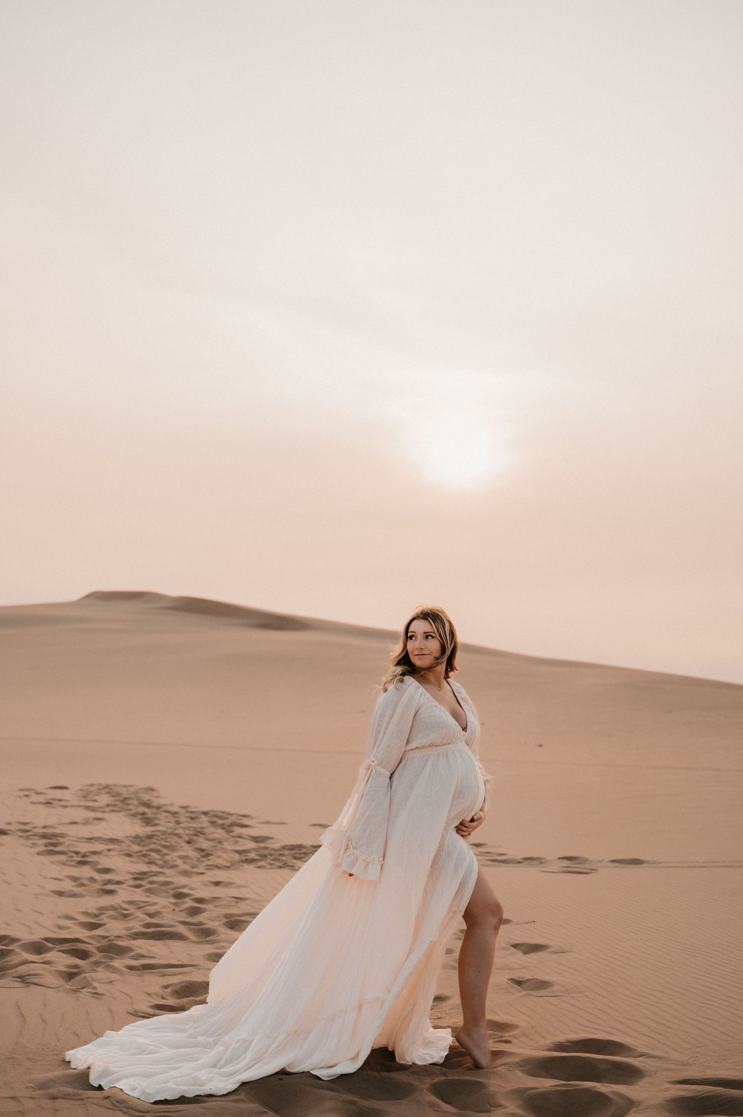 Maternity photo of woman in flowing white dress posed on desert sand dune.