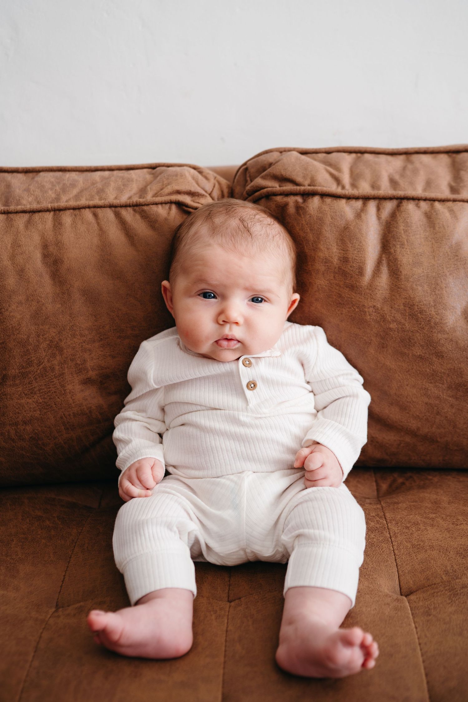 A baby wearing a white ribbed outfit sits on a brown leather couch showing bare feet.