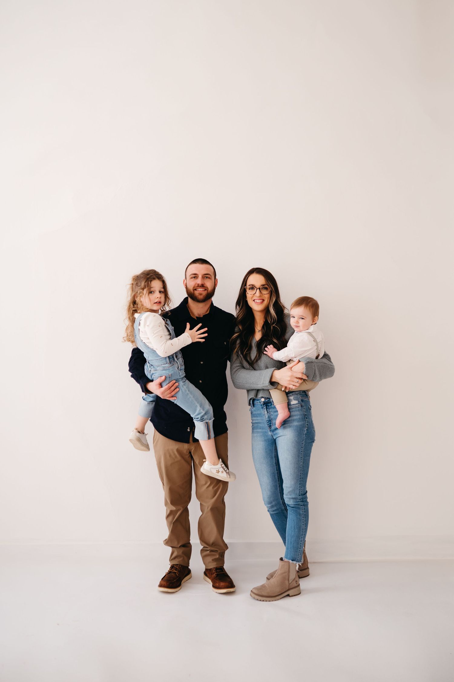 A family of four stands together against a white background wearing casual clothing in a minimalist studio setting.