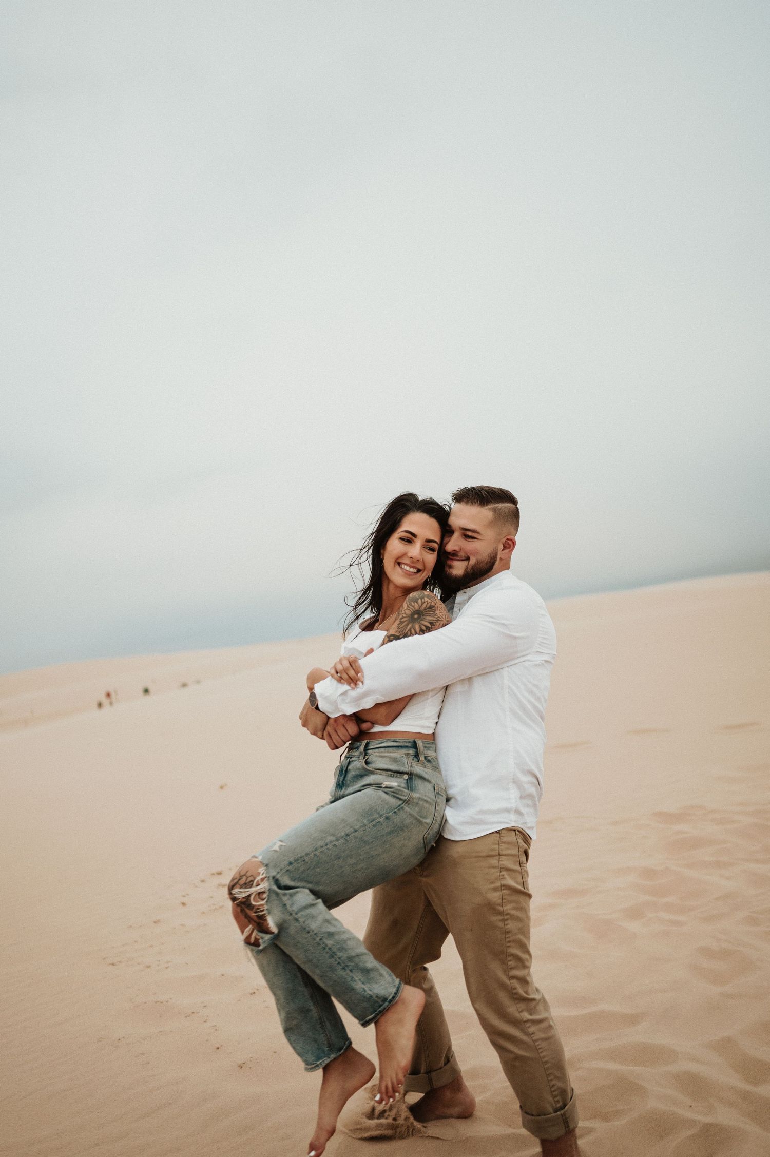 A couple shares a tender moment while standing on sand dunes against a cloudy sky.