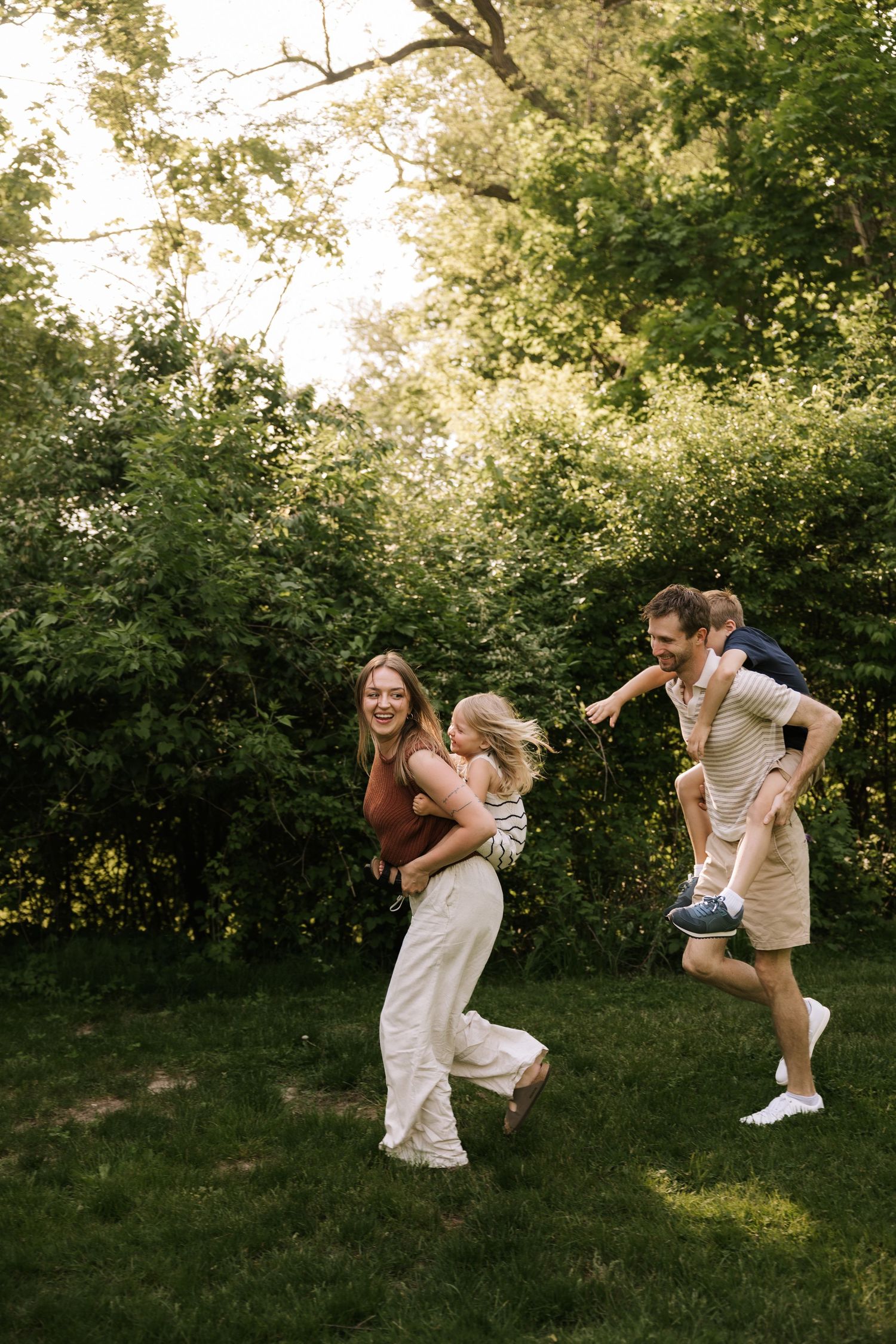 Family playing piggyback rides outdoors on green grass, laughing and enjoying a sunny day surrounded by lush trees.