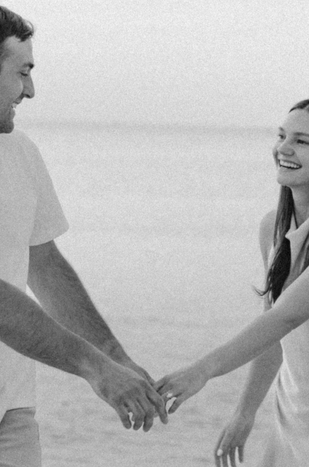 Black and white photo of a smiling couple holding hands on a beach, sharing a joyful romantic moment.
